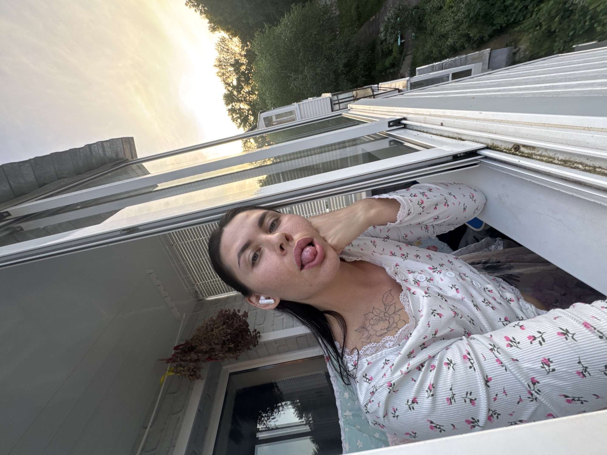 A young woman with dark hair, wearing a white shirt with pink floral print and white lace trim, is leaning out of an open window on a balcony. She has a tattoo on her chest and is wearing white earbuds. Her tongue is sticking out, and she is looking at the camera. Behind her, a bundle of dried herbs hangs from the ceiling. The balcony overlooks a green, wooded area with some buildings visible in the distance. The sky is a pale yellow, suggesting either sunrise or sunset. The scene appears to be in a residential area in Chaikovsky, Russia.