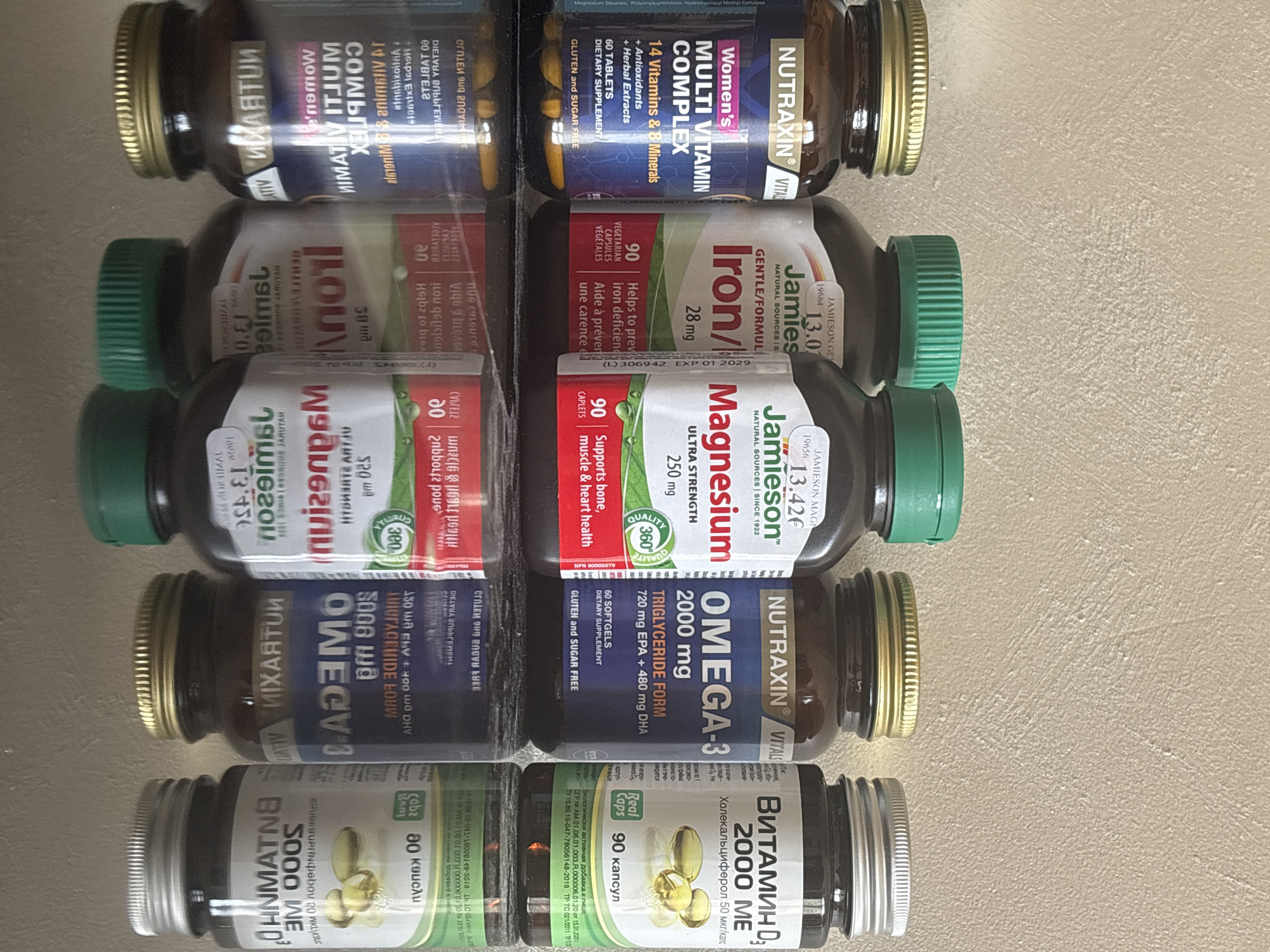 The image shows a close-up view of five bottles of vitamins and supplements, neatly arranged in a row. The first bottle from the top is "Nutraxin Vital Women's Multi Vitamin Complex." The second is "Jamieson Iron Gentle Formula". The third bottle is "Jamieson Magnesium Ultra Strength". The fourth bottle is "Nutraxin Omega-3". The last bottle from the bottom is "Витамин Д3" (Vitamin D3). All of the labels contain a mixture of Cyrillic and Latin script, and the labels appear to be Russian in origin, suggesting a Russian or Slavic community in the area. The background is a light beige color with a slight texture, possibly a countertop or shelf. The image appears to be taken indoors with neutral lighting. There is no indication that the location is in the city of Bar, Montenegro.