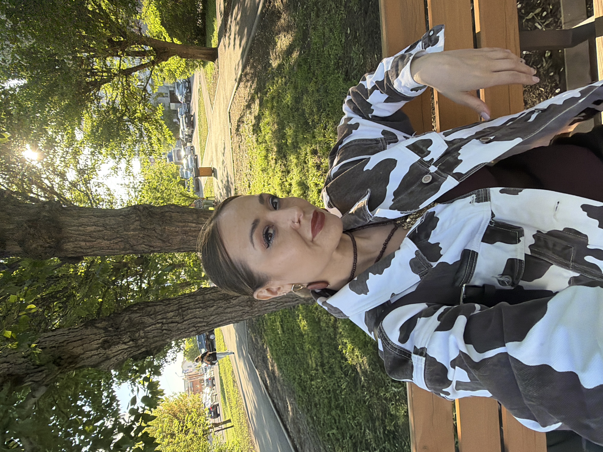 A woman sits on a wooden bench in a park, possibly in Krasnoyarsk, Russia, based on visual indicators. The background features a tree-lined street with parked cars and building details that hint at Eastern European or Russian urban architecture. The woman wears a cow-print jacket and jewelry. She is looking at the camera. The setting is outdoors, with natural light filtering through the trees. A paved walkway and grassy areas are also present.