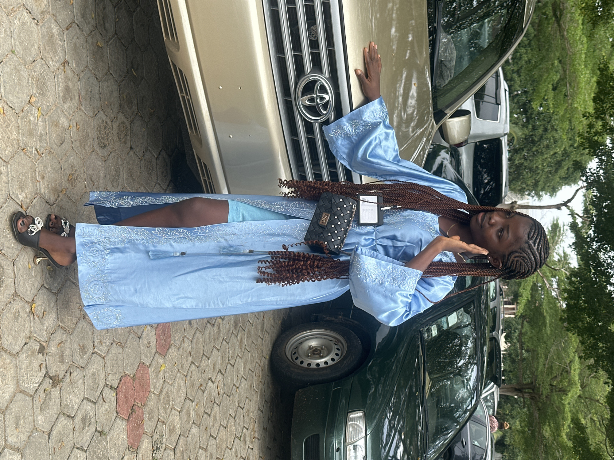 A young woman with braided dark hair stands in a paved outdoor area, possibly a parking lot, in Itoga, Nigeria. She is wearing a flowing, light blue abaya with intricate white embroidery along the sleeves and hem. The abaya is open at the front, revealing a shorter blue garment underneath. She is also wearing black heeled sandals with silver embellishments.
The woman has a black quilted purse with silver studs hanging from her left shoulder. She is looking to her right, with her left hand raised to her face, as if shielding her eyes or gesturing.
In the background, two cars are visible. To the left, a gold-colored SUV, possibly a Toyota, is parked. To the right, a dark green sedan is parked. Trees with green foliage are visible behind the cars and above them, suggesting an outdoor, possibly semi-urban or suburban environment. The ground is paved with hexagonal concrete stones. The overall lighting suggests it is daytime.