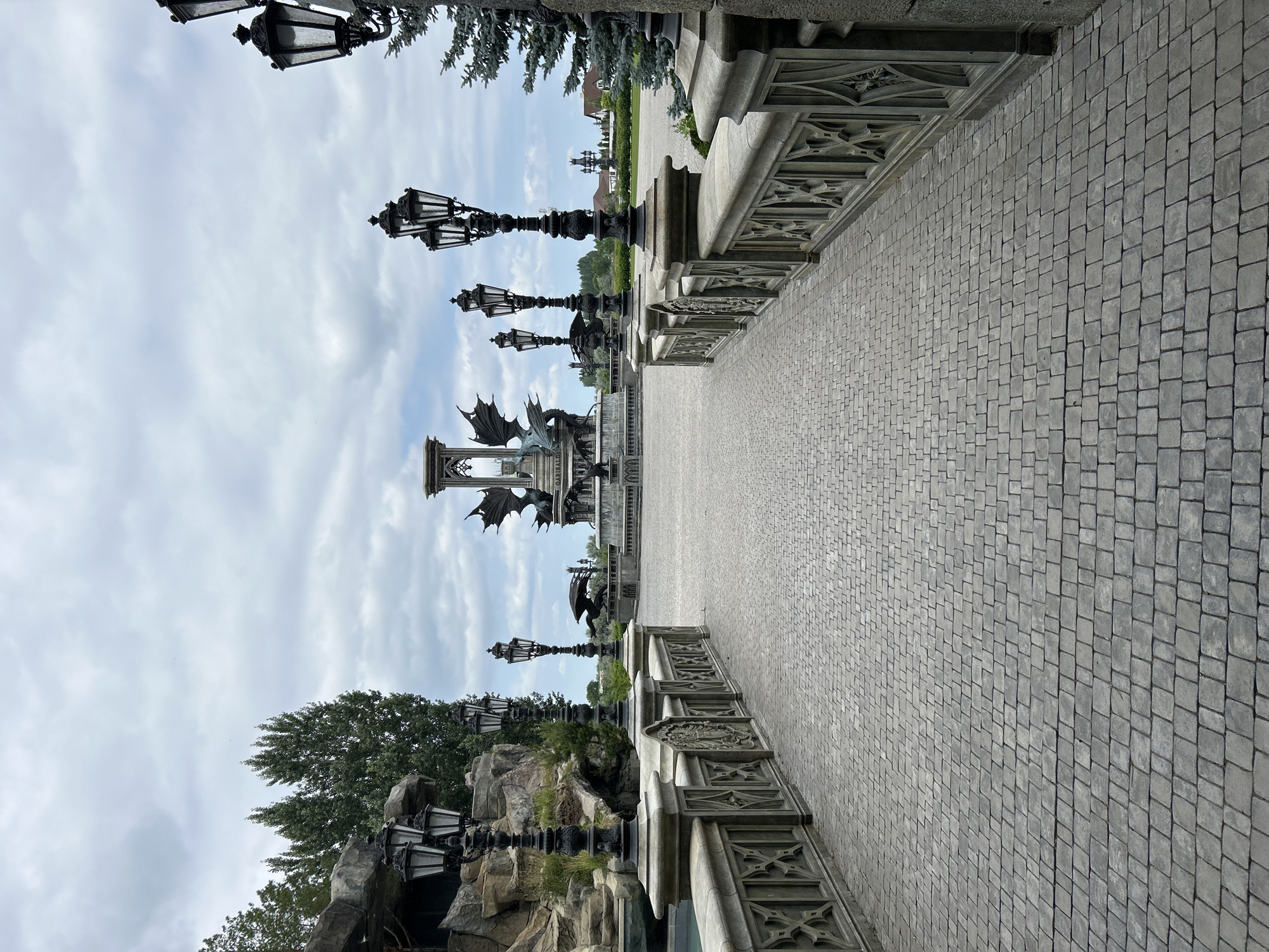 A paved walkway leads toward a large, ornate fountain featuring dragon sculptures. The walkway is lined with decorative lampposts and stone railings. The fountain is centrally located in an open area with trees and buildings visible in the background under a cloudy sky. The scene appears to be an outdoor public space, possibly a park or attraction.
