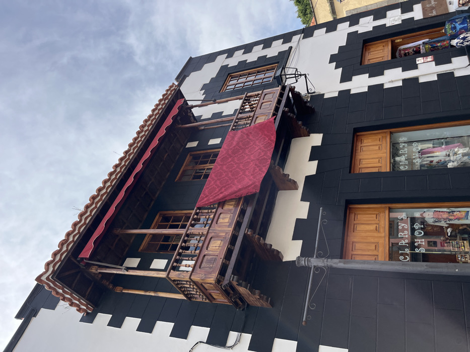 The image depicts a two-story building with a black and white painted exterior, located in Teror, Spain at coordinates 28.06, -15.547. The building has a prominent balcony extending out from the front of the second floor, which is made of wood and painted brown. There is a red awning over the balcony and another, larger red fabric draped over the balcony's railing, hanging down to the first floor. The balcony has a wooden railing and a metal streetlamp. The building has two storefront windows, one on each side of the balcony, both displaying the name "Clarry's Shop" in white letters on a black background. The windows are made of glass and have wood frames. The lower level of the building appears to be a commercial storefront. The scene is taken from a street-level perspective, looking up at the building. The sky is cloudy and the weather appears to be mild.