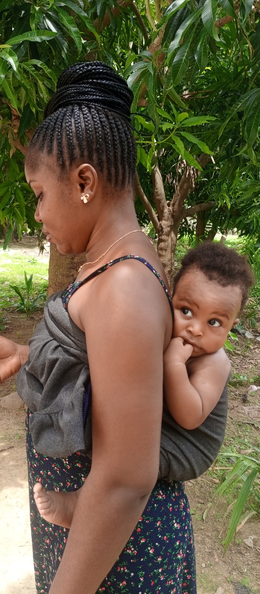 In an outdoor setting in Keffi, Nigeria, a young woman is carrying a baby on her back, secured with a gray cloth. The woman has dark skin, braided hair styled in a bun, a gold necklace, and floral stud earrings. She is wearing a dark dress with a floral pattern. The baby, who also has dark skin, is looking to the side and has a finger in its mouth. Green foliage and tree trunks are visible in the background.