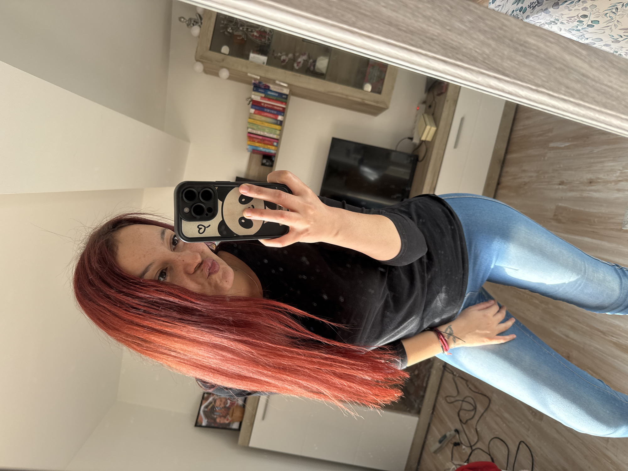 A woman is taking a selfie in a mirror. She has red hair, freckles, and is wearing a black shirt and blue jeans. The phone case is panda-themed. The reflection shows a TV, books, a cabinet, and part of a bed. The floor is light brown wood laminate. The location cannot be determined as Varna, Bulgaria, without additional information.
