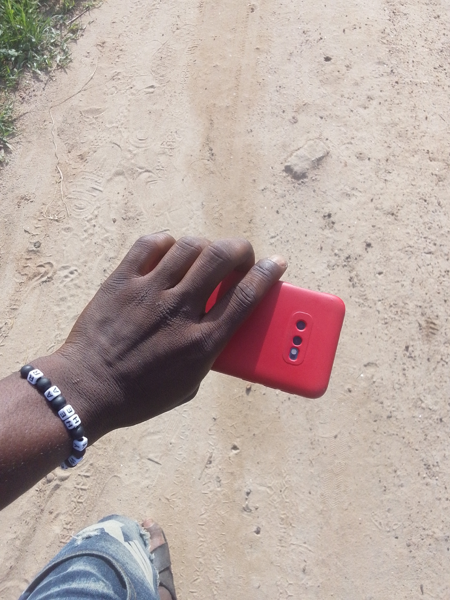 The image depicts an overhead view of a dark-skinned individual holding a red smartphone while standing on a dirt path in Keffi, Nigeria. The hand is positioned centrally, holding the red smartphone horizontally with the back facing upwards, revealing a vertical triple-camera cutout on the upper left side. A black beaded bracelet with white square letter beads is worn on the left wrist; visible letters include 'R', 'E', 'A', 'V', 'E', 'N', 'H', 'E'. Part of a lower leg is visible in the bottom left, clad in distressed blue denim jeans, and a dark sandal is partially seen on the foot. The ground is composed of light brown, dry soil or sand, marked with numerous shoe prints, tire tracks, and scattered small rocks and debris. Green vegetation is visible along the upper left edge of the path.
