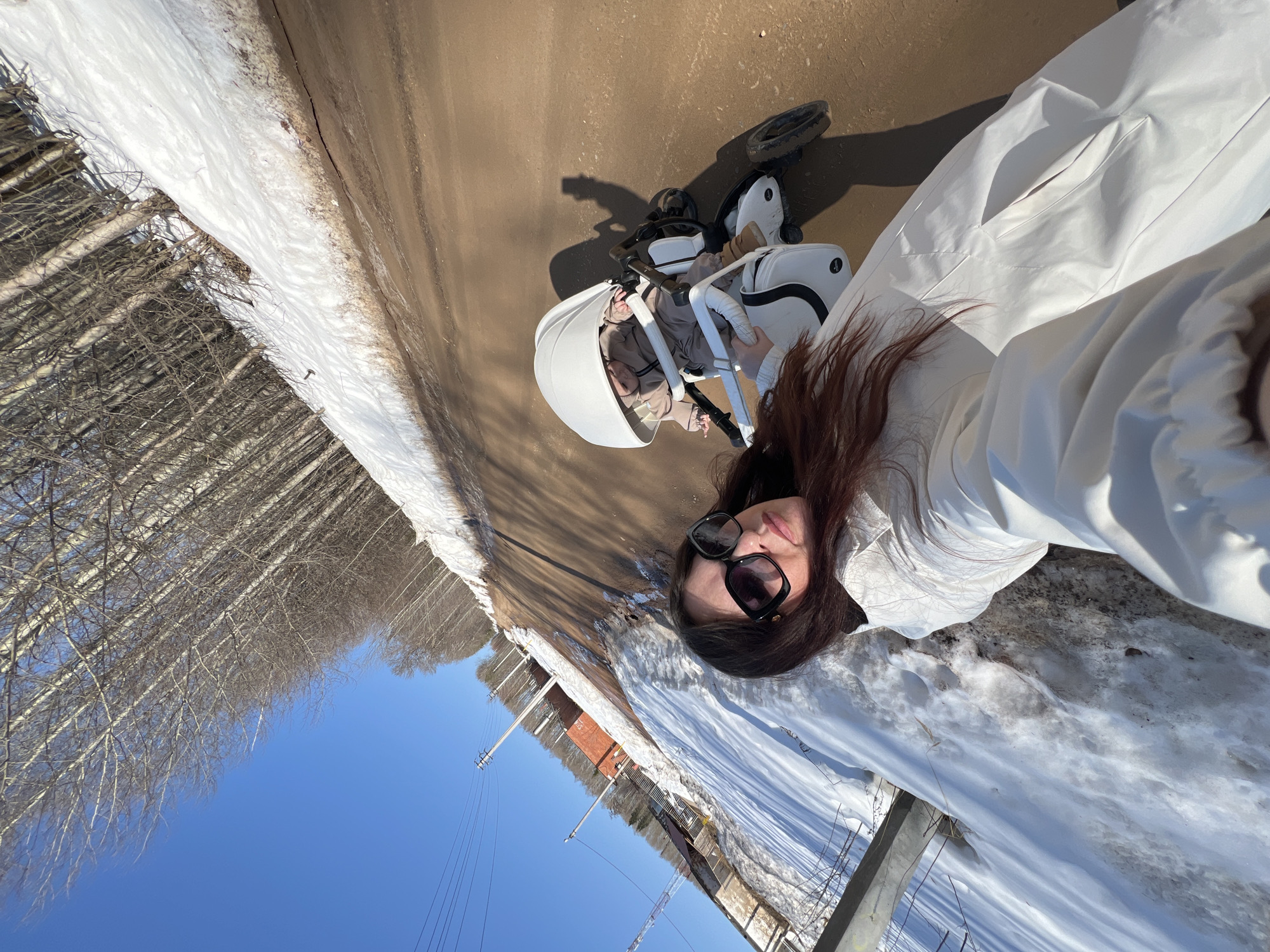 A woman wearing sunglasses and a white jumpsuit is taking a selfie on a road, with a baby in a white stroller. To the left of the road is snow and trees. On the other side of the road, near the bottom, is a brick building. The sky is blue. The photo may have been taken in Noviy, Russia.