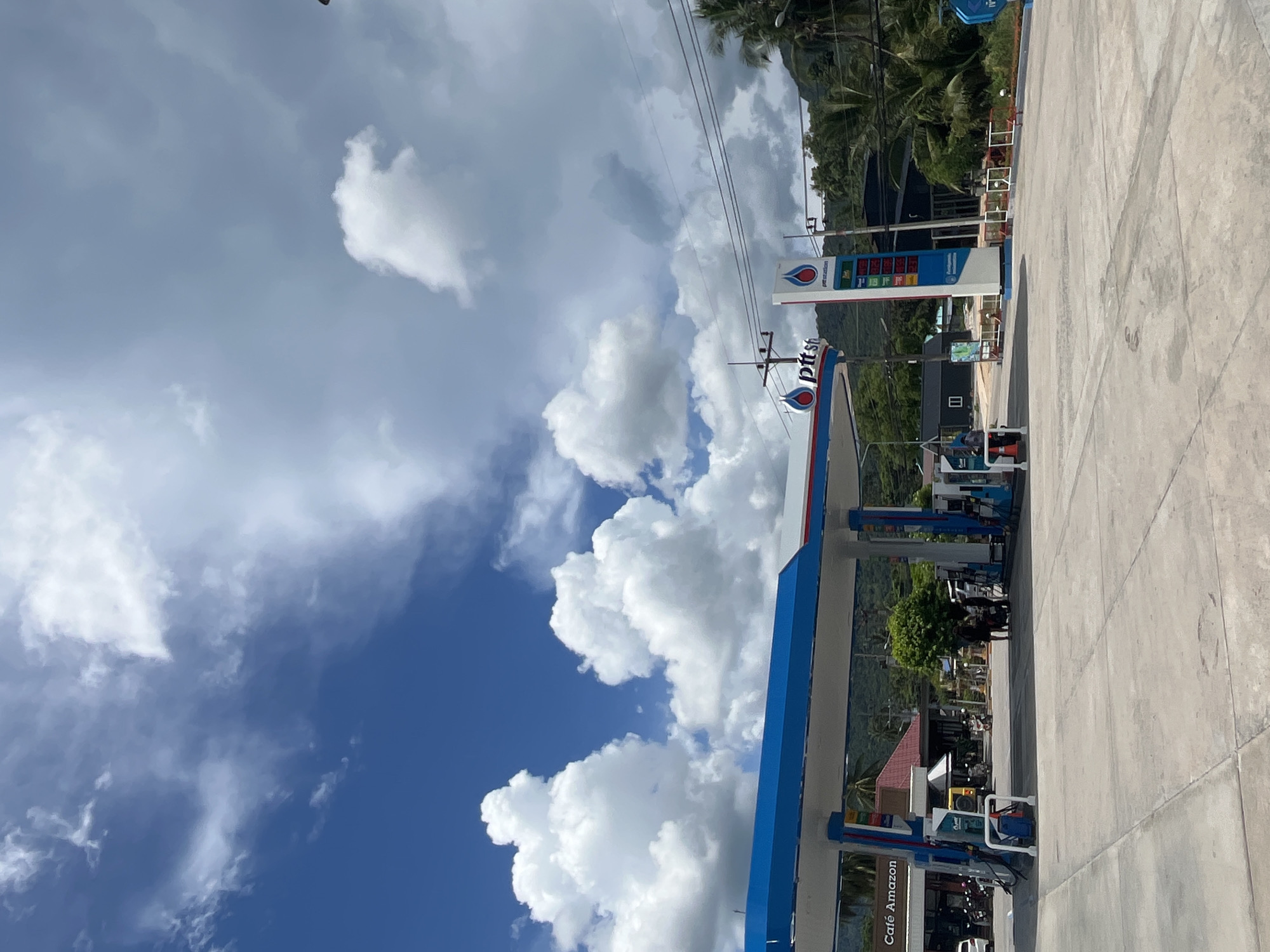 A gas station with a PTT station branded canopy is visible in the foreground under a bright, cloudy blue sky. The canopy is supported by white pillars and has blue trim. To the left of the canopy, a building with a sign for "Café Amazon" is visible. Several motorcycles are parked in front of the buildings. A tall PTT station sign displaying fuel prices is located to the right of the canopy. In the background, lush green hills and palm trees are present. The ground is paved with concrete. Several people are standing near the gas pumps. The scene depicts a typical day at a gas station in a tropical location.