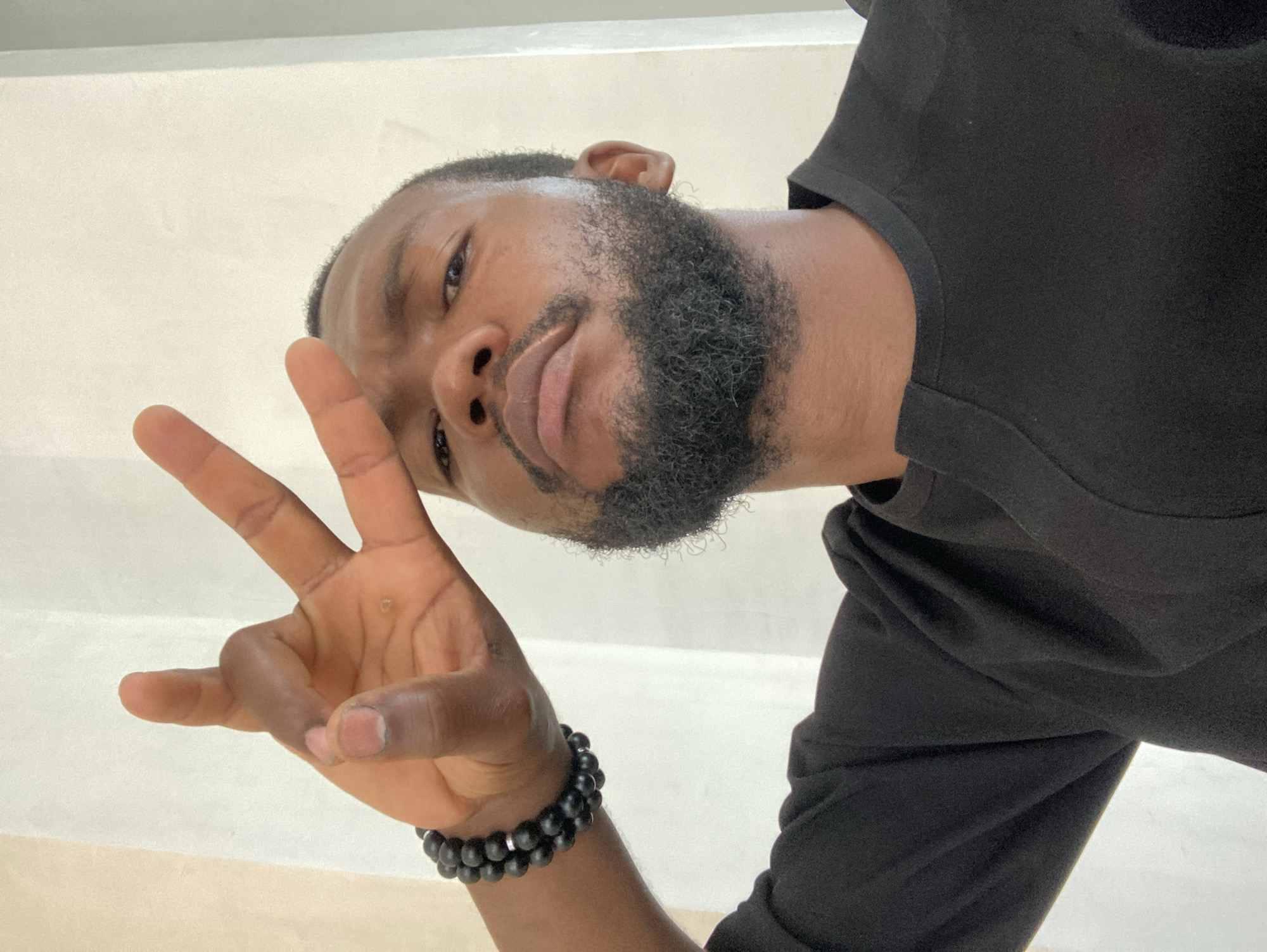 A low-angle close-up shows a dark-skinned person with a black beard and short hair. They are wearing a black shirt and a beaded bracelet on their left wrist. The person is holding up their left hand, making the "ok" gesture. The background appears to be an off-white wall or surface. The image's location is unverifiable with the available information.