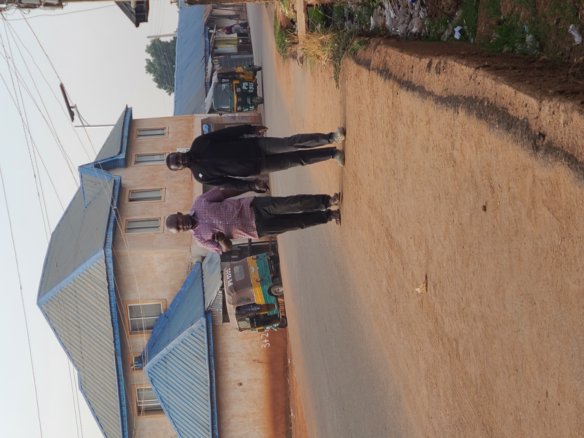 The photograph captures a street scene in Jos, Nigeria, at ground level with the camera oriented to the side. Two men, both Black, are standing side-by-side on a dirt road that runs through a residential area. One man wears a checkered shirt and the other is dressed in a black jacket. Buildings with corrugated metal roofs line the street. In the background, a small three-wheeled motorized vehicle known as a "keke" or "tricycle" is parked. The edge of the road on the right side is slightly elevated with some trash and foliage visible on the higher ground. Utility wires crisscross overhead.