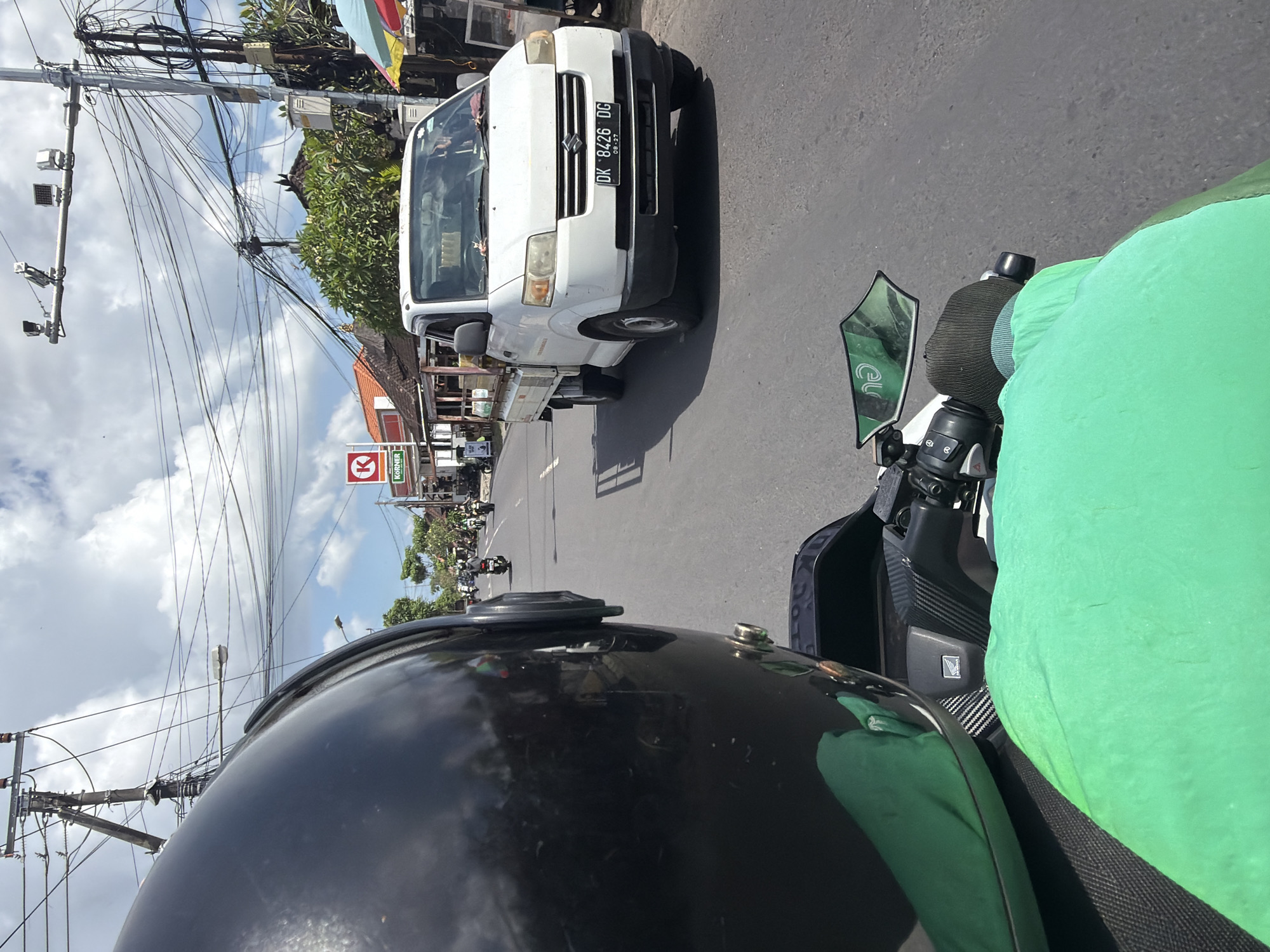 The image, taken from a motorbike in motion, captures a street scene in North Kuta, Indonesia. A white Suzuki Carry pick-up truck with license plate "DK 8426 DG" can be seen to the right. The motorbike rider, visible via their black helmet and green clothing, is positioned behind the handlebars and rearview mirror. In the background, a "KORNER" convenience store sign and overhead power lines are visible. Motorbikes are travelling on the street. The weather appears to be sunny with some clouds.