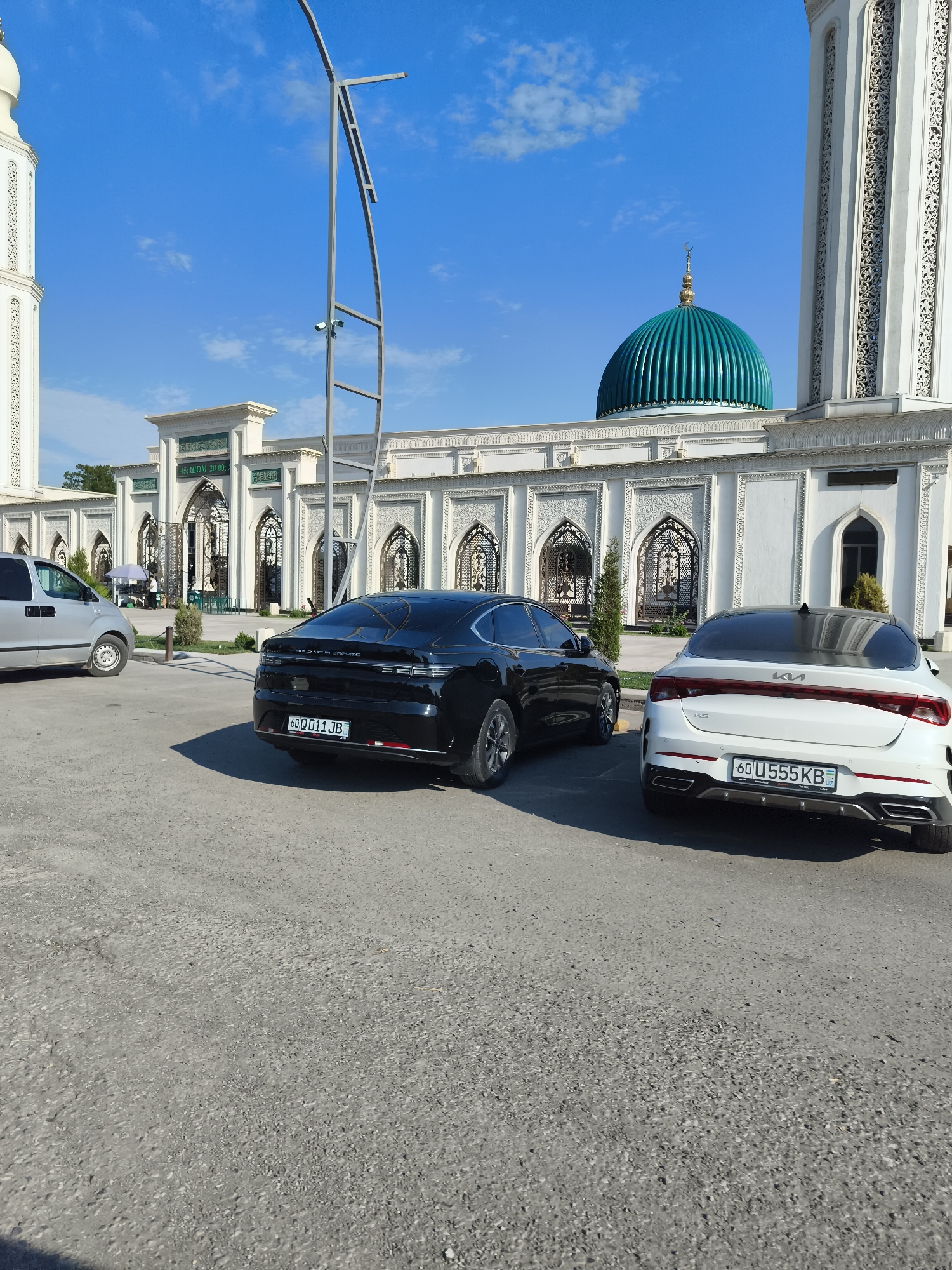 The image shows the exterior of the Azam-o-Rafiq Mosque in the city of Buz, Uzbekistan, on a clear, sunny day. In the foreground, two cars are parked on an asphalt lot. The car on the left is a black sedan with license plate "60 Q 011 JB". The car on the right is a white Kia sedan with license plate "60 U 555 KB". Behind the cars is the mosque's facade, featuring white walls with intricate decorative patterns and arched doorways. A large, teal-colored dome is visible atop the mosque, topped with a crescent moon. A tall, white minaret is also part of the mosque's architecture, located to the left of the dome. A modern-looking street lamp structure stands in the middle ground. In the background, a few trees and other structures are partially visible. The sky is bright blue with a few wispy clouds.