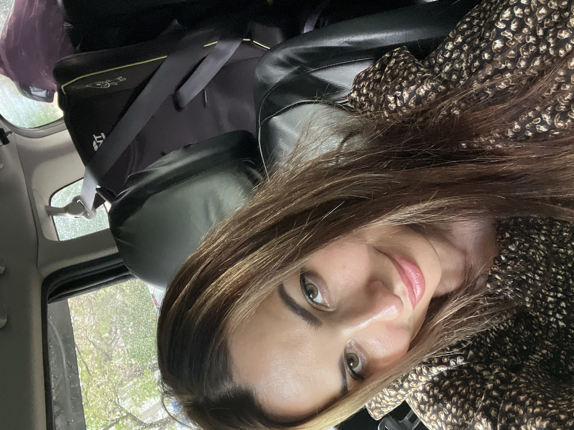 A selfie of a woman taken from inside a vehicle. The woman, with long brown hair and light skin, is looking at the camera with a slight smile. She is wearing a brown and black patterned top. The background shows the interior of the car, including a child car seat and a window with rain streaks. The scene is likely in an urban environment, given the presence of trees and buildings visible through the window.