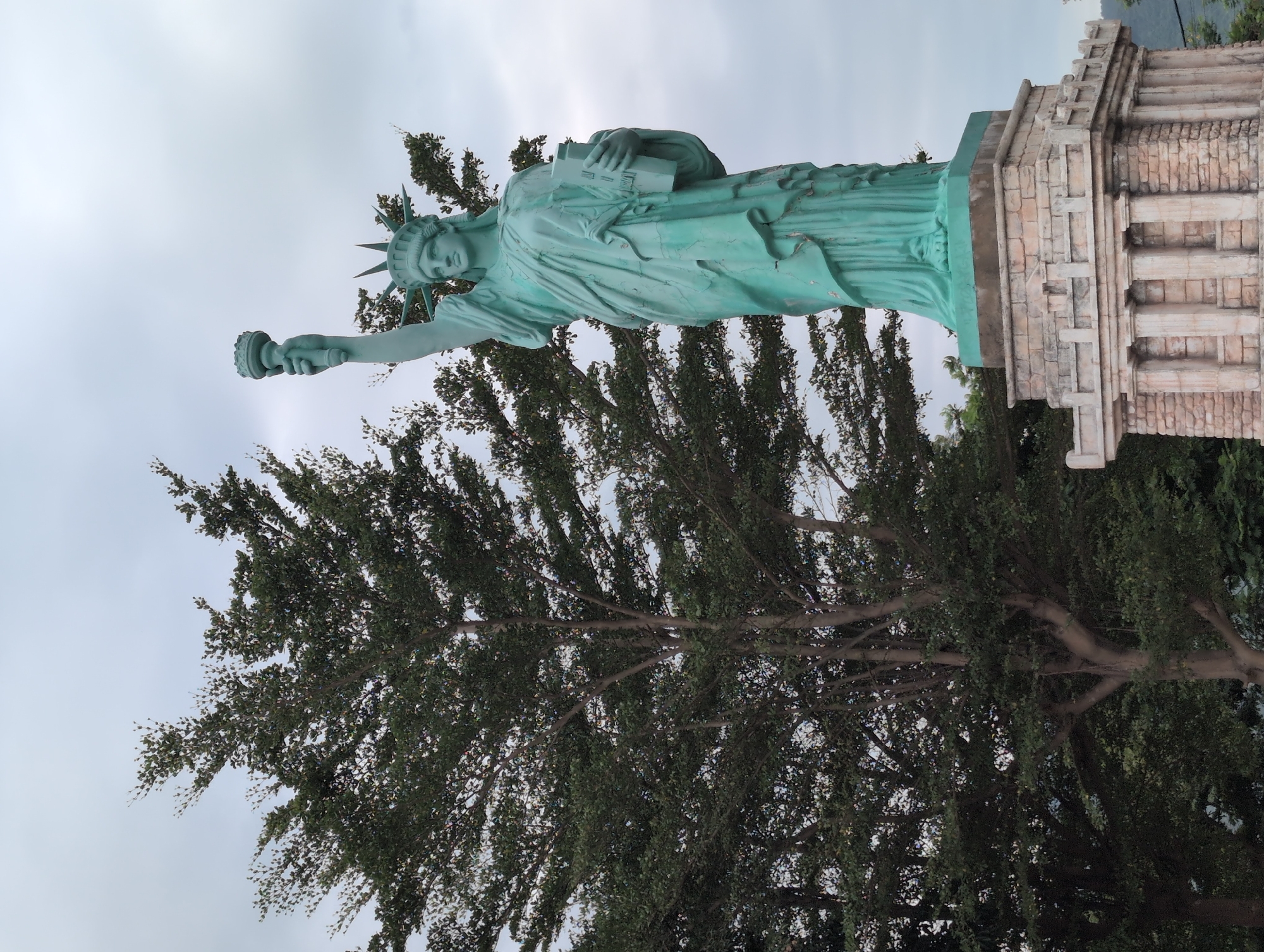 The image shows a replica of the Statue of Liberty, a copper-colored statue painted green, standing on a stone pedestal. The statue is positioned to the right of a large tree with dense foliage, and both are set against a cloudy sky. The base of the statue is a multi-layered structure made of stone blocks and columns, reminiscent of classical architecture. The Statue of Liberty replica is located in Da Nang, Vietnam, specifically in the Sun World Danang Wonders amusement park. The scene is an outdoor setting with a focus on the statue and the surrounding vegetation.