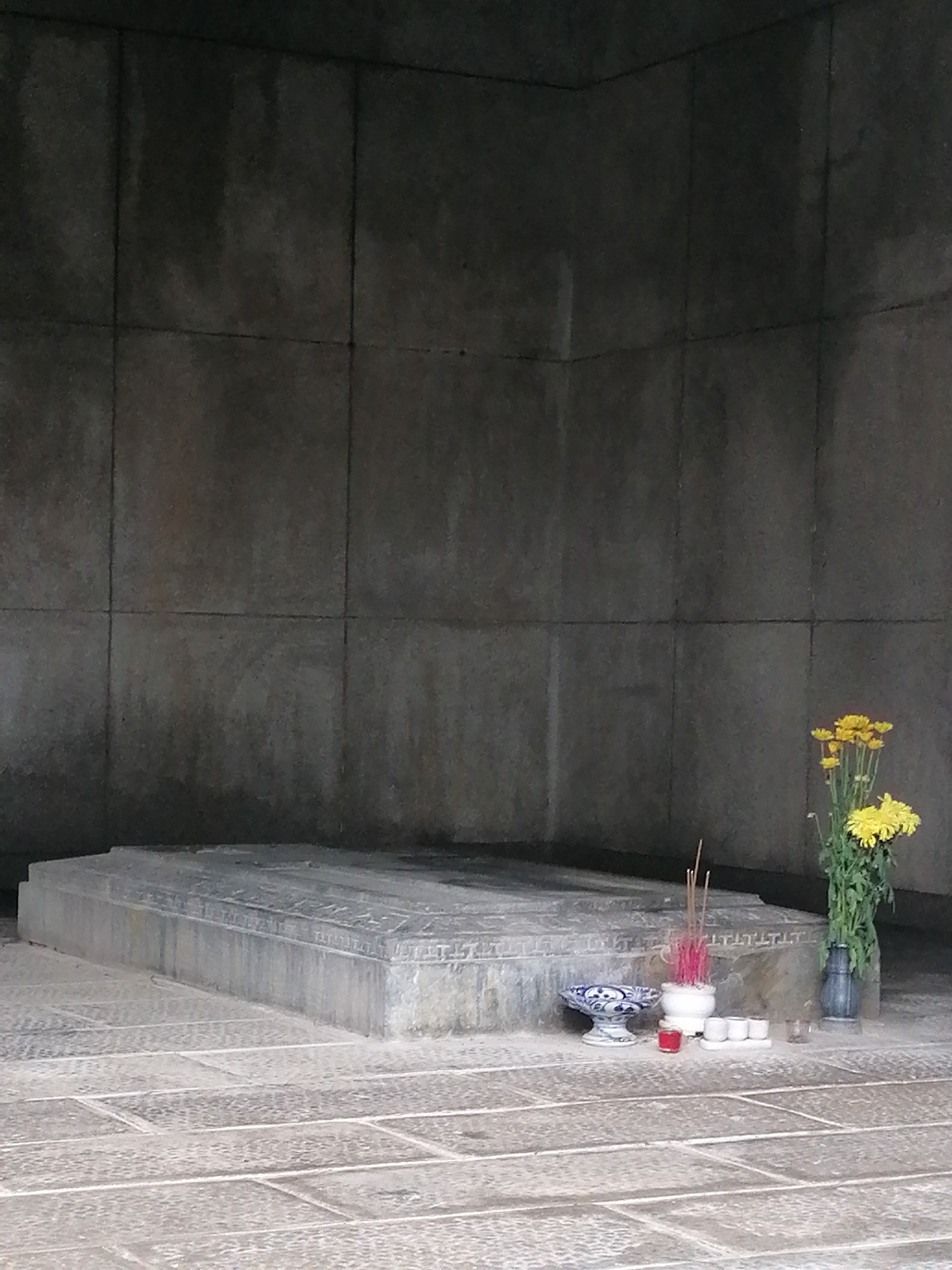 The image captures a solemn interior space, likely a tomb or memorial shrine, characterized by stark stone architecture and offerings.
In the foreground, a large, rectangular stone structure, resembling a sarcophagus or a substantial altar, occupies the left side. It is crafted from grey stone with carved details along its edges and top surface. The floor beneath and around it is paved with matching grey stone tiles.
To the right of this stone structure, an arrangement of offerings is placed on the tiled floor. This includes a vibrant bouquet of yellow chrysanthemums held in a dark vase, several slender red incense sticks standing upright in a small white holder, a distinctive blue and white patterned ceramic bowl, a single red candle, and three small white cups, possibly for liquid offerings.
The background consists of dark grey or almost black stone slab walls, meeting in a corner and extending upwards. These walls show signs of weathering, with vertical streaks and variations in tone, adding to the ancient and somber ambiance.
The lighting throughout the scene is subdued, creating a quiet and contemplative atmosphere. There are no people visible, but the presence of fresh flowers, incense, and other offerings indicates a recent act of remembrance, respect, or worship. Given the context of Cam Ly, Đà Lạt, Vietnam, this setting is consistent with a traditional Vietnamese memorial or tomb site. No visible text is present in the image.