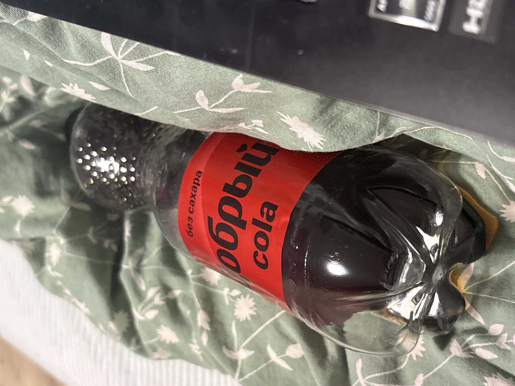 A partially visible bottle of "Добрый Cola без сахара" (Good Cola without sugar) is lying on a green fabric with a white floral pattern. Part of a dark-colored object, possibly electronic, is visible in the top right corner. The scene is located in Прикамский, Russia, based on the bottle's branding.