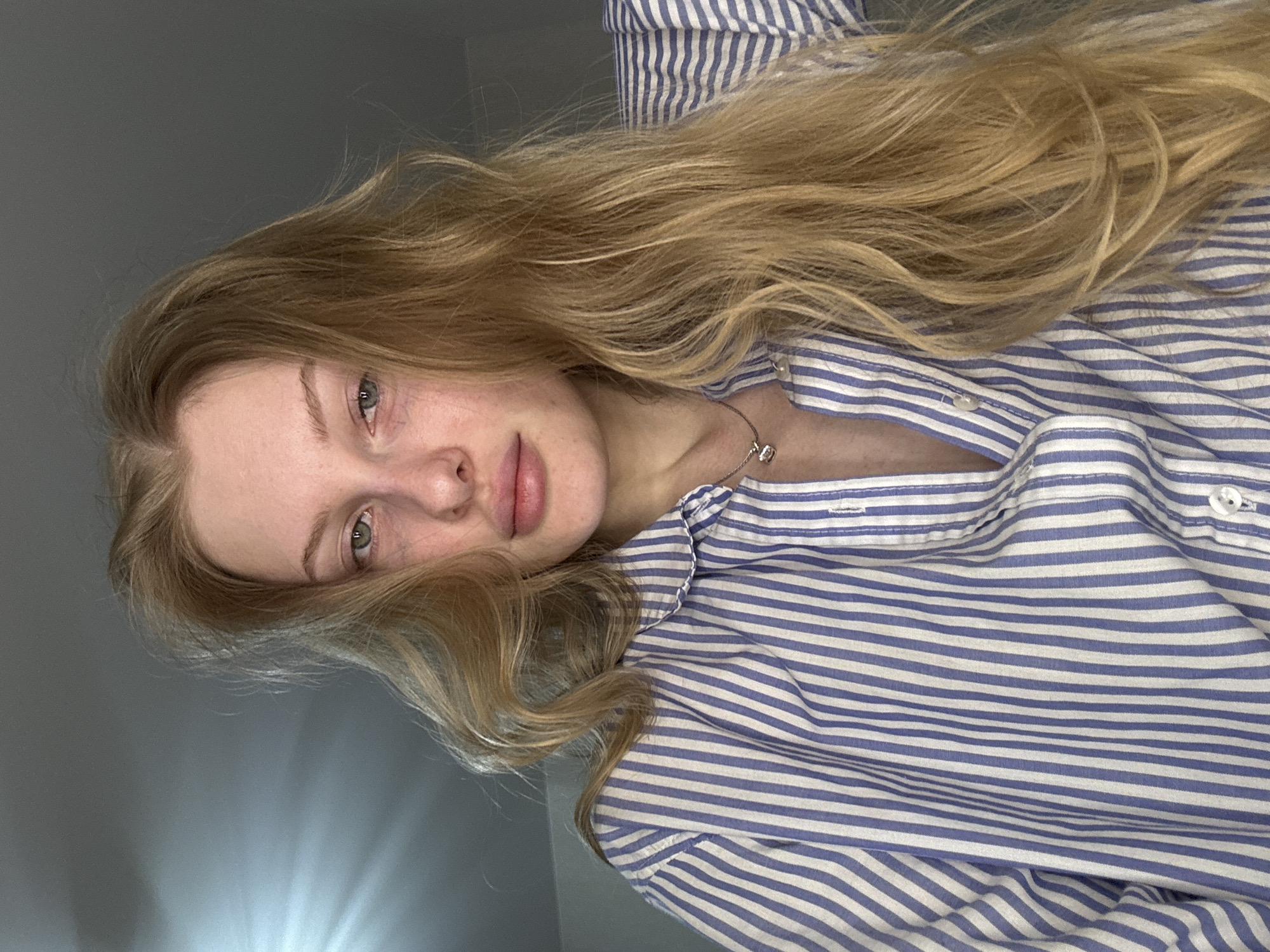 A fair-skinned young woman with long blonde hair is wearing a blue and white striped button-down shirt. She is facing the camera with a neutral expression. A silver necklace is visible on her neck. The background is a plain gray wall. The image appears to have been taken indoors. The location cannot be determined from the image.