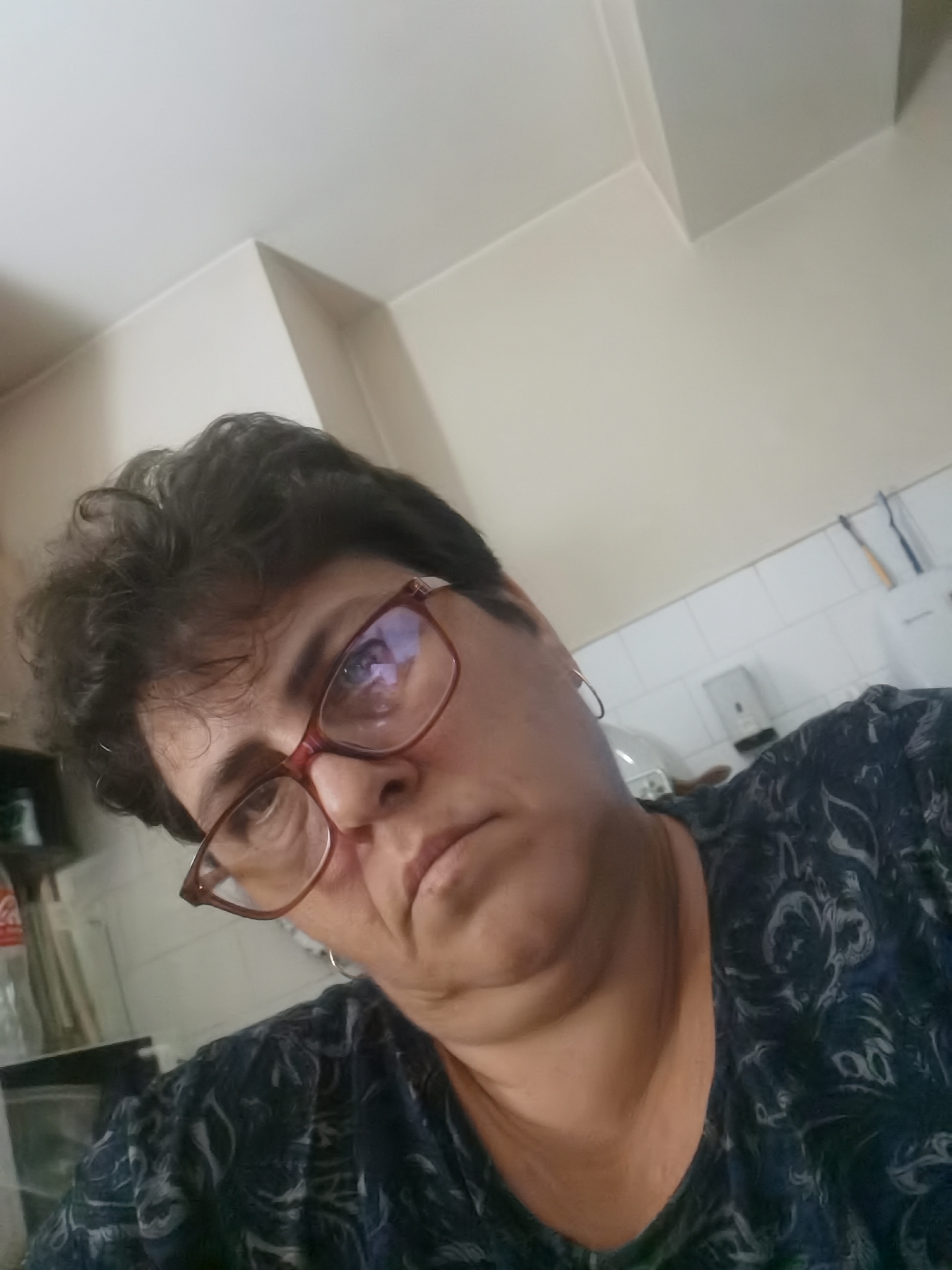 The image is a selfie taken by a woman wearing glasses and a dark, patterned shirt. She has short, dark, curly hair and is looking down and to her left. The background appears to be an indoor setting, possibly a kitchen or bathroom, with white tiled walls and a white ceiling. There are some objects visible in the background, including what looks like a bottle and some cleaning supplies. The lighting is artificial. The photo's metadata or context does not indicate any specific location in Lomets, Bulgaria.