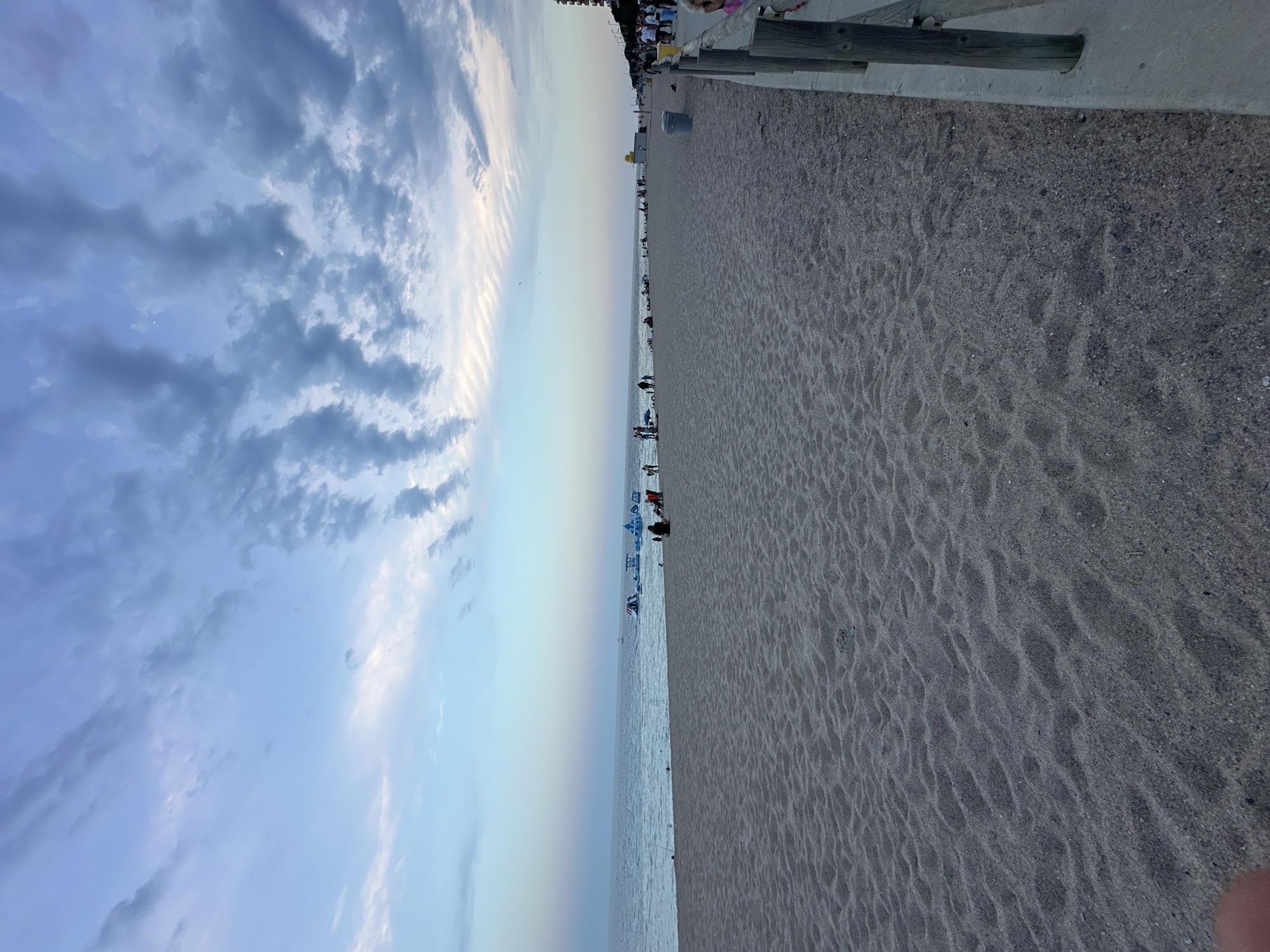 The image captures a wide view of a sandy beach in Blanes, Spain, during the late afternoon or early evening. The sky is a soft gradient of blue and white, with scattered clouds creating a textured pattern. The ocean stretches out to the horizon, appearing calm and reflecting the muted sky colors.
On the beach, several people are visible, scattered across the sand. Some are seated, while others appear to be walking or standing. In the distance, offshore, there are inflatable water attractions, indicating a recreational area. The sand itself is fine-grained, with visible undulations and footprints, suggesting recent activity.
To the right of the beach, a wooden boardwalk or promenade is delineated by wooden posts and a concrete edge. This area appears to be more developed, with some individuals walking along it, and a yellow structure that could be a kiosk or a public amenity. Further along the promenade, buildings and possibly more people are visible. The overall atmosphere is relaxed, characteristic of a beach setting at the end of the day.