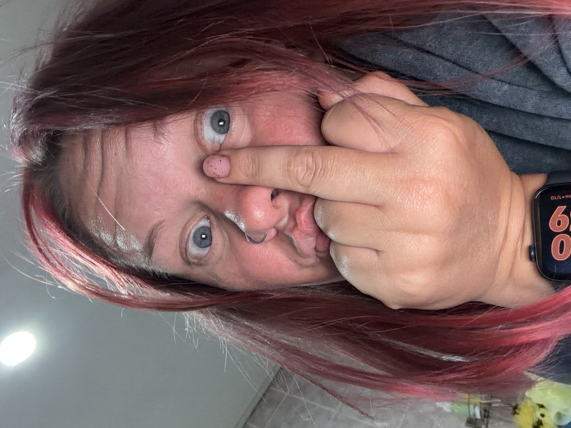 A close-up selfie shows a woman with pink-red hair, light skin, and blue-grey eyes. She has a silver nose ring and her lips are puckered. Her right hand is partially obscuring her face, with a finger touching the side of her nose near her eye. The nail on that finger is painted light pink with dark speckles. She is wearing a dark grey shirt and a black smartwatch is visible on her left wrist, displaying digital numbers. The background consists of a white wall with a light source casting a bright reflection. The location and city of Chaikovsky, Russia cannot be determined from the image content.