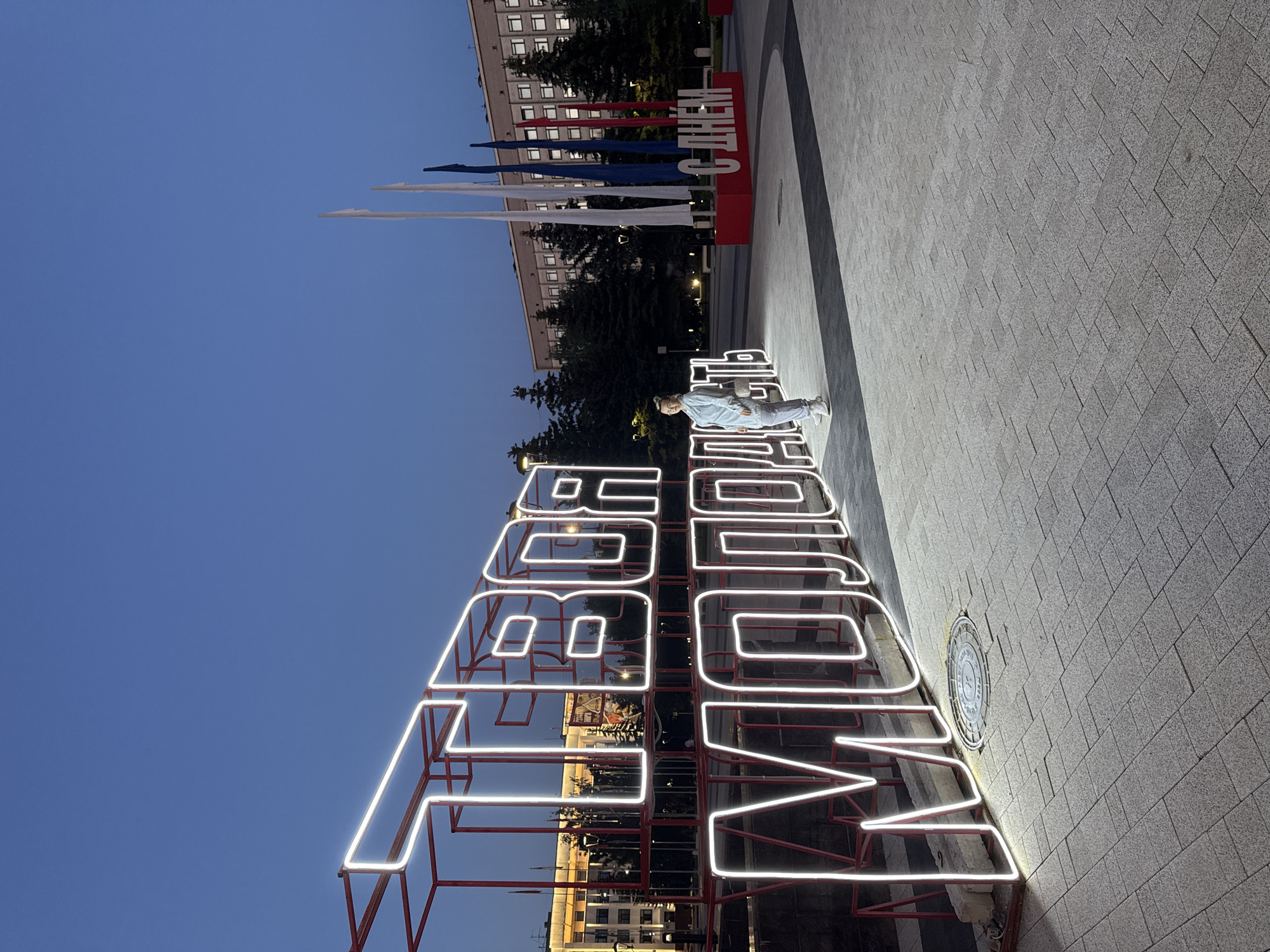An evening outdoor shot captures a public square in Vologda, Russia, at dusk. The central feature is a large, freestanding sign spelling "ВОЛОГДА" (VOLOGDA) in Russian, constructed from a red metal frame brightly illuminated with white neon lights. A person, dressed in light-colored casual attire, stands to the right of this illuminated sign, looking towards the viewer.
In the background, a large red sign with white lettering partially reads "С ДНЕМ" (C DNEM), suggesting it is part of a "Happy Victory Day" message. Behind this sign, several tall flagpoles display long banners in red, white, and blue, echoing the colors of the Russian flag. Further back, multi-story buildings with illuminated windows and dark trees are visible against the deep blue twilight sky. The ground is covered with grey rectangular paving stones, and a circular manhole cover is visible near the base of the "VOLOGDA" sign. The scene is well-lit by the sign's neon glow and ambient urban lighting, creating a clear and serene atmosphere.