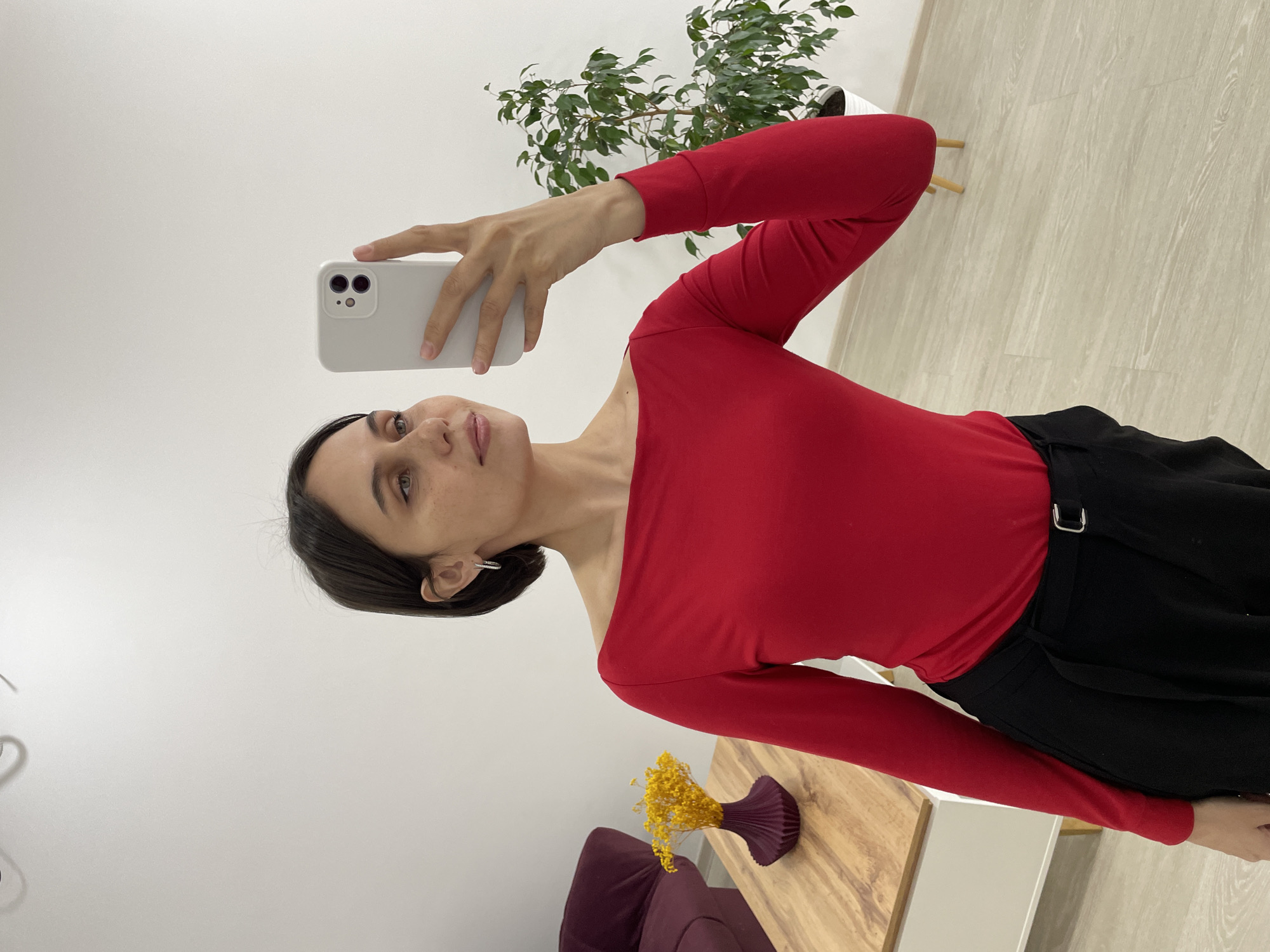 A young woman is captured in a mirror selfie indoors, holding a white smartphone with a light-colored case in her right hand. She has dark hair, light eyes, and scattered freckles across her face, with a neutral to slightly soft expression. She is wearing a long-sleeved, bright red top, possibly off-the-shoulder on her left, paired with black trousers or a skirt that features a silver buckle. A simple hoop earring is visible in her left ear.
The setting appears to be a well-lit, modern interior with plain white walls and a light-colored wooden floor. In the background to her right, a lush green potted plant sits in a white pot. To her left, part of a small wooden table or shelf is visible, holding a unique purple ribbed vase filled with dried yellow flowers. A section of a dark-colored (possibly burgundy or purple) armchair or sofa can be seen near the table. The overall atmosphere suggests a clean and contemporary space, potentially in Izhevsk, Russia. The bright, even lighting indicates it is likely daytime or an artificially well-lit environment. No discernible text is visible in the image.