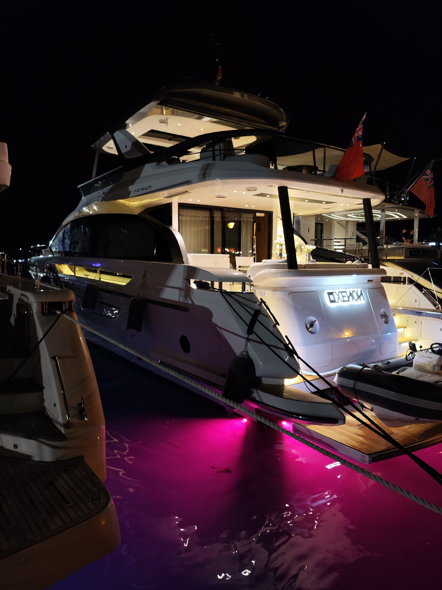 The image depicts a large white yacht docked at night in Budva, Montenegro. The yacht is illuminated by interior lights and accented by pink underwater lights that cast a vibrant glow on the water. The yacht features multiple decks, sleek lines, and is branded with "AZIMUT" on its side and "HXEMOM" on its stern. A smaller inflatable dinghy is tied to the stern of the yacht, and other moored boats are visible in the background. The dark night sky serves as the backdrop, with the illuminated yacht and its reflection in the water being the central focus. The scene suggests a luxurious and tranquil evening at a marina.