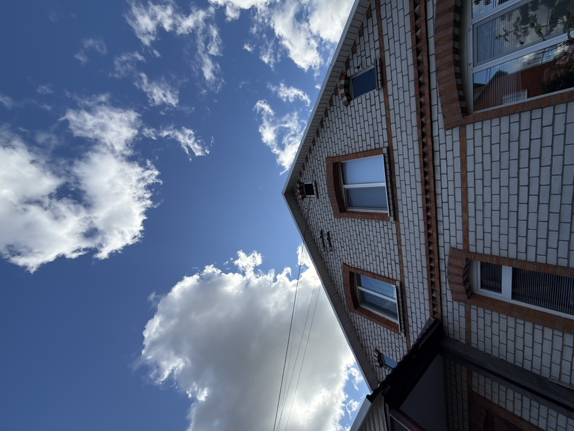 The image is a low-angle, outdoor shot of the facade of a brick house against a blue sky with scattered white clouds. The house has a pitched roof and appears to be two stories tall. It is constructed of light-colored bricks with red brick accents around the windows and along a decorative band below the upper windows. There are several windows visible, some with blinds. Overhead, power lines stretch across the frame. The overall impression is of a residential building on a clear day. The prompt states the location is Zheleznogorsk, Russia. There are no people or animals visible in the image, and no actions are taking place.