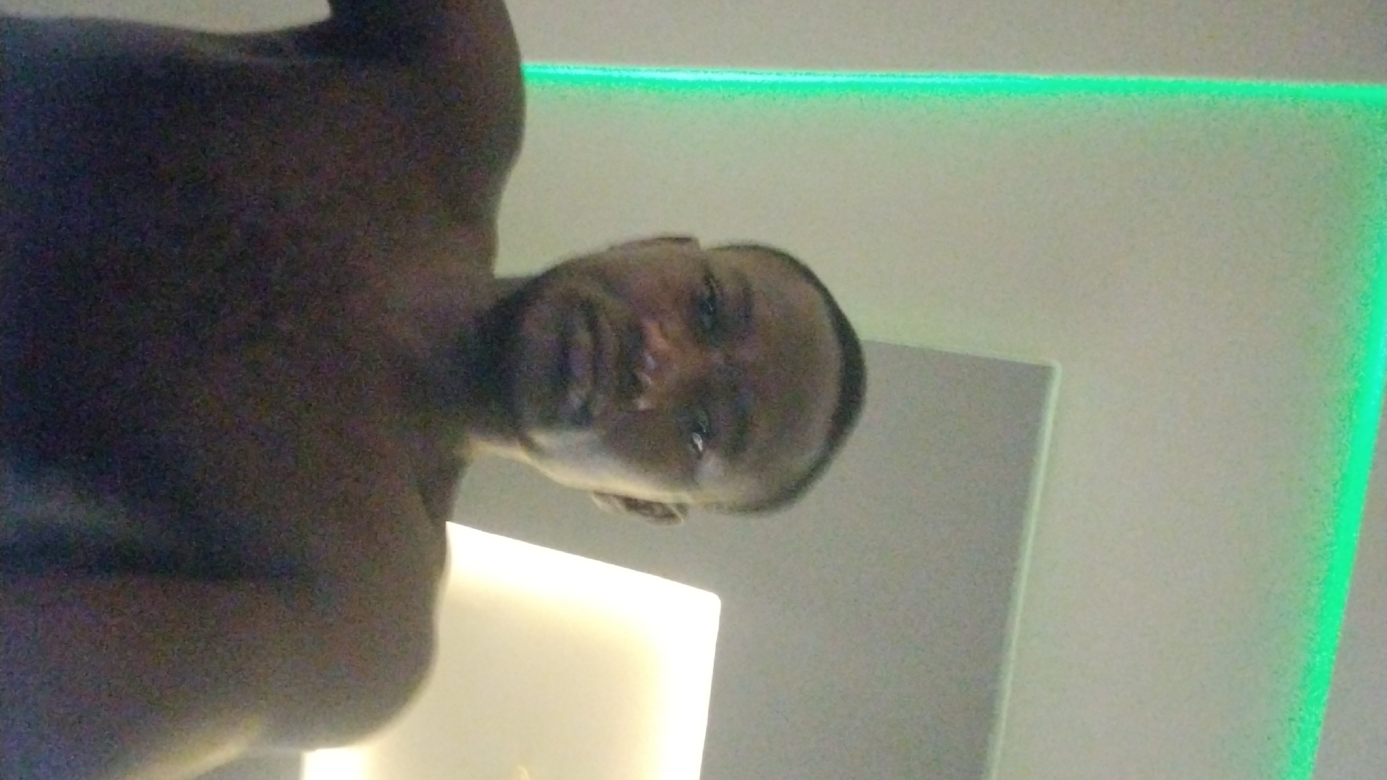 The image shows a man's upper torso and head. He has dark skin and short hair. The background is a white or light-colored wall with a green light strip along the top edge. In the lower portion, there appears to be a white, rectangular light source. The photo is taken from an unusual angle, looking upwards at the subject. No specific location within Jos, Nigeria, is identifiable from the image.