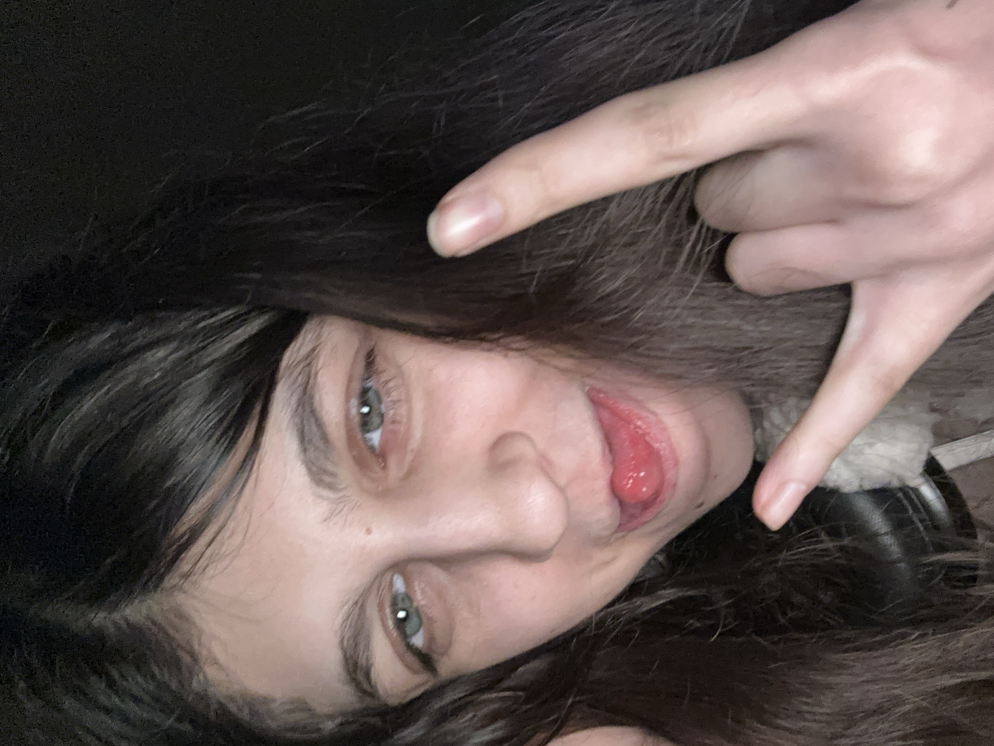 A close-up, rotated selfie features an individual with long, dark hair, captured indoors in what is contextually known as Moscow, Russia. The person looks directly at the viewer with an expressive gaze from their light-colored eyes, and their tongue is slightly extended in a playful manner. One hand is raised, forming a "rock on" or "horns" gesture with the index and pinky fingers extended, while the other fingers are curled. The background is dark and out of focus, suggesting low-light conditions or an indoor setting at night. A subtle detail of what appears to be a furry collar or a part of a headphone band is visible near the person's right ear. The overall impression is one of casualness and playfulness. There is no visible text in the image.