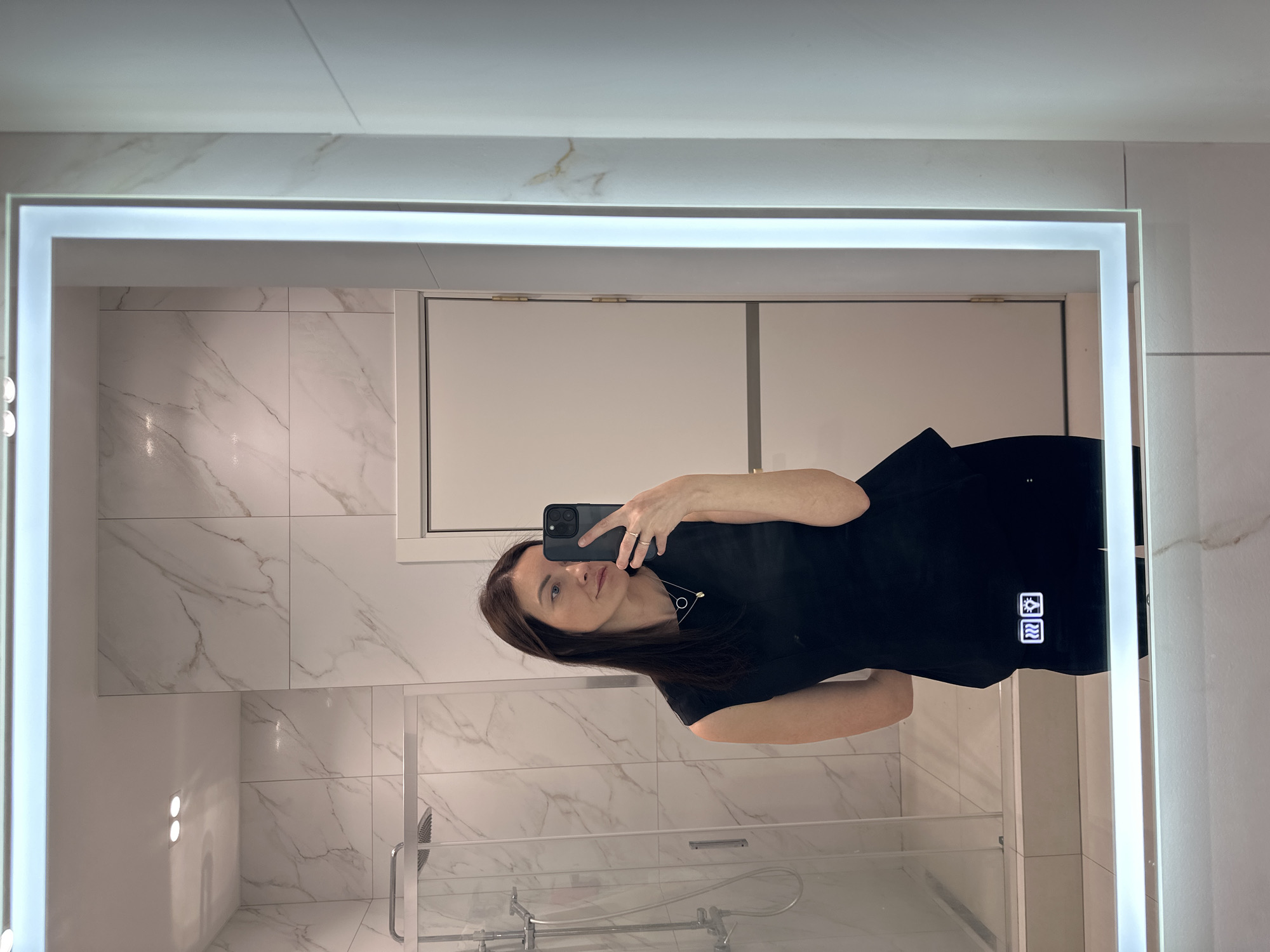 A woman is taking a selfie in a bathroom mirror. The bathroom features white marble tile walls and a shower. The mirror is rectangular with a light border. The woman is wearing a black top and holding her phone in her right hand.