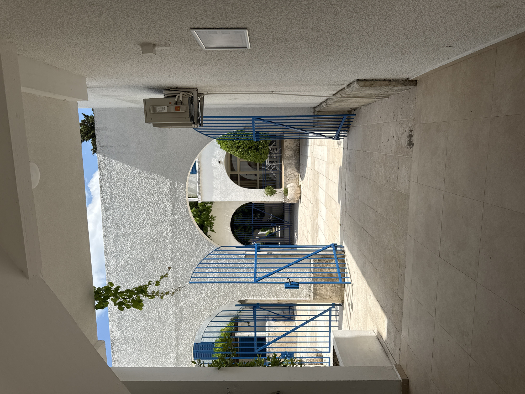 This media file depicts a sunlit outdoor walkway or courtyard, likely in Boreti, Montenegro. The scene is dominated by white, textured walls and beige tiled flooring. The walkway is bordered by a blue metal railing and gates, suggesting a secure or private area.
Several architectural features are prominent, including arched openings that reveal glimpses of greenery and a blue-shuttered window. One archway has a blue metal gate that appears to lead to a different section of the property or a building interior. Above one of the gates, an air conditioning unit is mounted on the white wall, indicating a temperate climate where cooling is needed. A light fixture is visible on the textured ceiling of the walkway.
A small, leafy vine is growing up a section of the white wall, adding a touch of natural color to the otherwise monochromatic architecture. The sunlight casts strong shadows across the tiled floor, suggesting it is daytime, possibly mid-morning or afternoon. The sky visible through an opening is blue with scattered white clouds, indicating fair weather.
There is no clear activity or event taking place; the scene appears to be a still moment in time, focusing on the architecture and ambiance of the location. No people are visible in the frame. A small, rectangular sign is affixed to the ceiling, but the text is not legible. The overall impression is one of a tranquil, possibly Mediterranean-style setting.