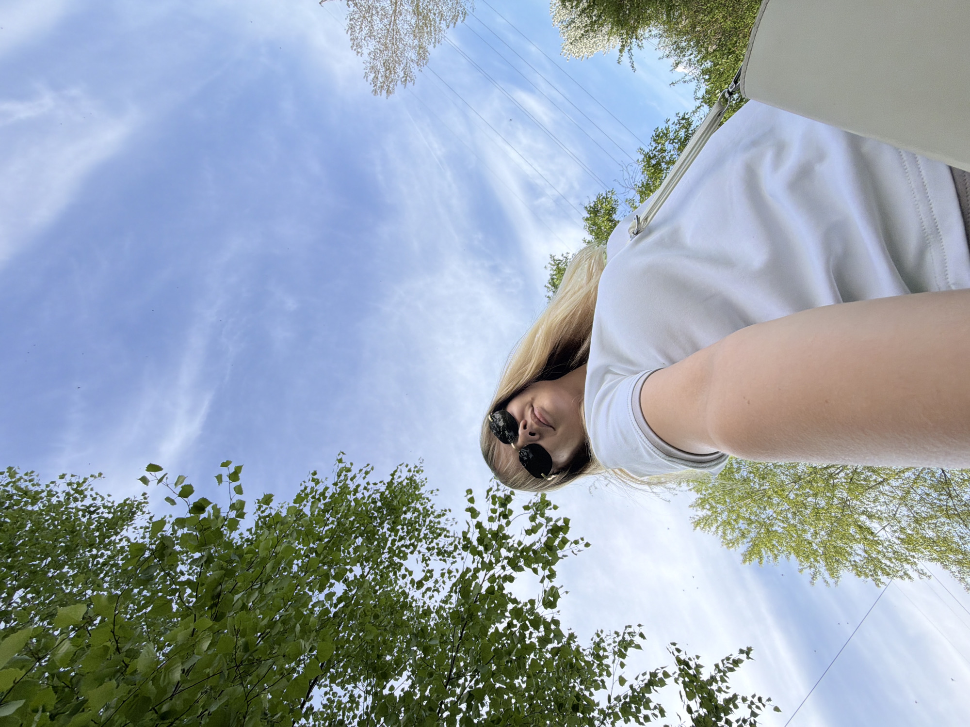 The image shows a low-angle view of a person wearing sunglasses and a white shirt, positioned against a backdrop of trees and a blue sky with wispy clouds. The person's face is tilted downwards, and they appear to be looking at the camera. Power lines are visible in the upper part of the frame. Based on the location metadata, the image was taken in Chaikovsky, Russia.