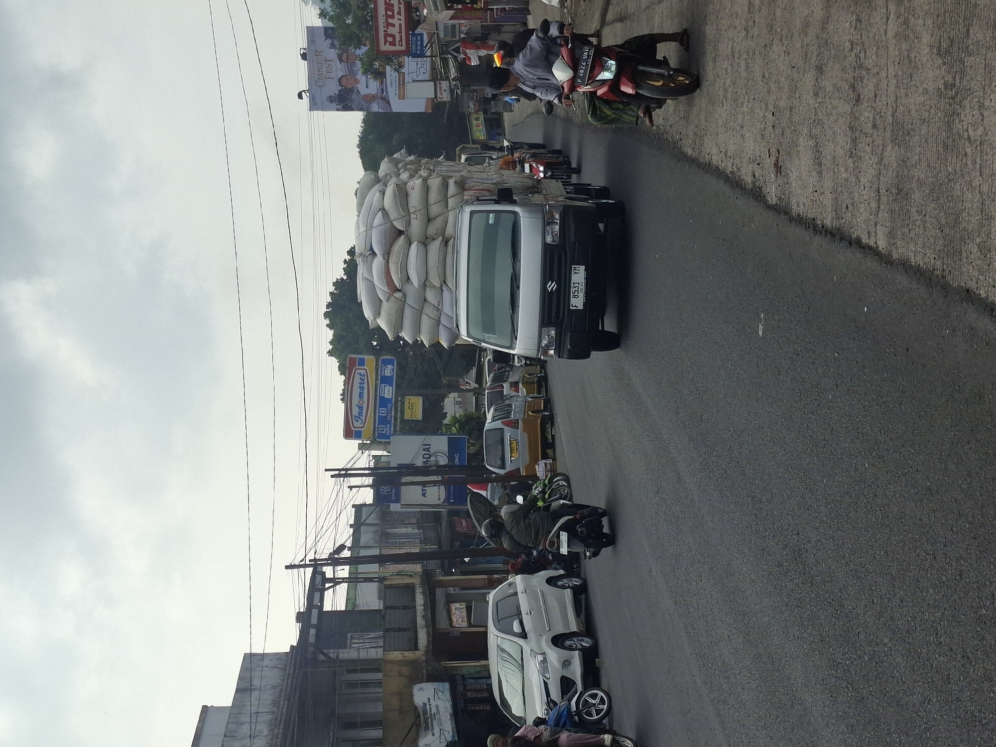 The image depicts a street scene in Gekbrong, Indonesia, on an overcast day. A silver Suzuki truck heavily loaded with stacked sacks of goods is the central focus, traveling on a paved road. The truck has a license plate reading "F 8531 YM". Several other vehicles are visible: a white minivan to the left, a yellow minivan behind the truck, and various motorcycles. People are seen riding motorcycles, including one individual in a grey shirt and green sarong on a red motorcycle to the right, and another on a dark motorcycle in the foreground. The roadside features buildings, signage for businesses like "Indomaret" and "D'TOP", and utility poles with overhead wires. The environment appears to be a commercial area with shops and a somewhat busy street.