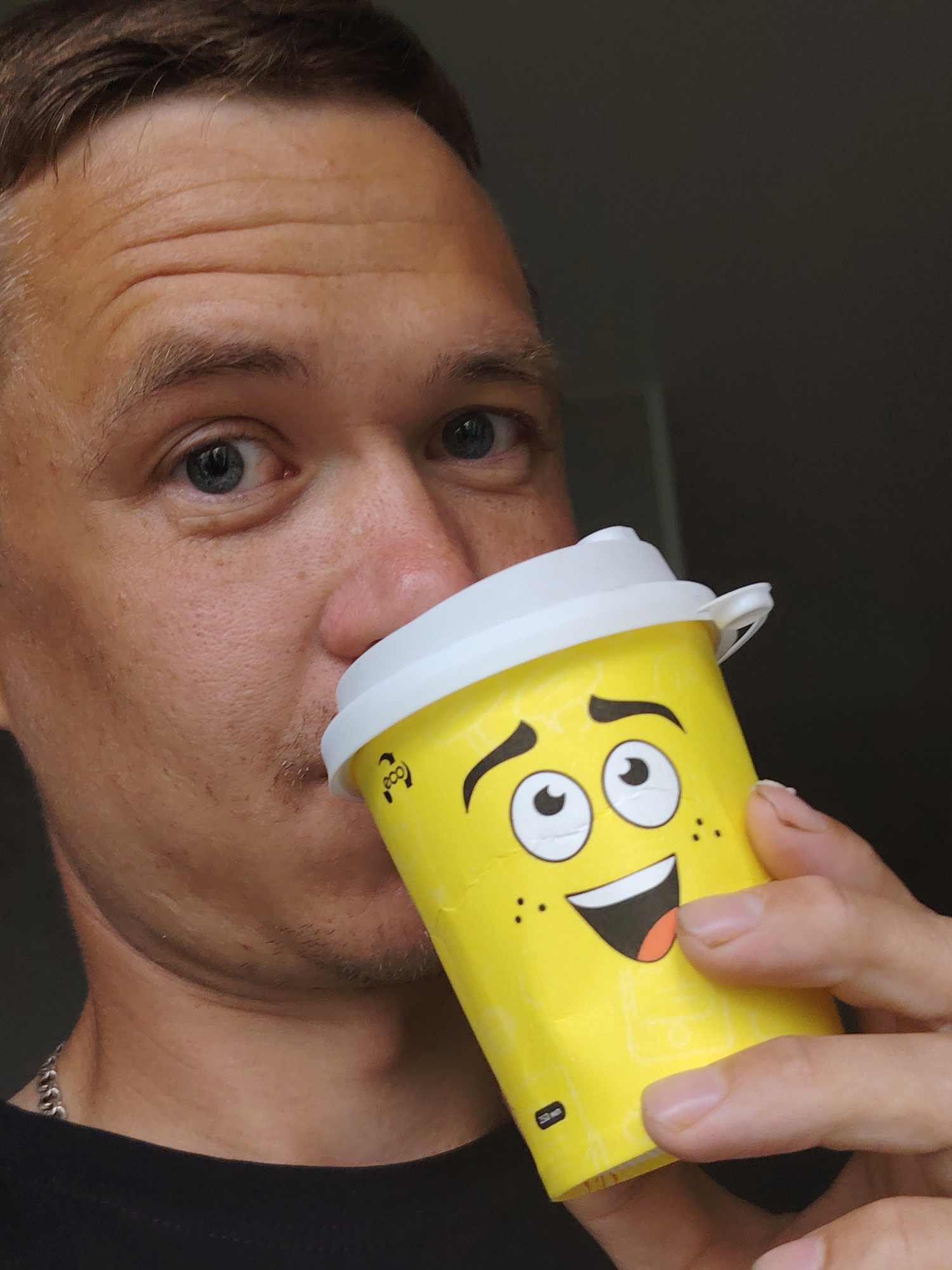 An extreme close-up shot shows a man drinking from a yellow disposable cup with a cartoon face. The man's face, neck, and chest are visible. He has short brown hair, blue eyes, and stubble on his chin and upper lip. He is wearing a black t-shirt and a silver chain necklace. The cup has a white lid and a smiling cartoon face with black eyebrows, white eyes with black pupils, and a wide, open mouth with an orange tongue. Small black dots are scattered on the cup's surface, resembling freckles. The background is dark and out of focus. The image was taken in the city of Chaikovsky, Russia.