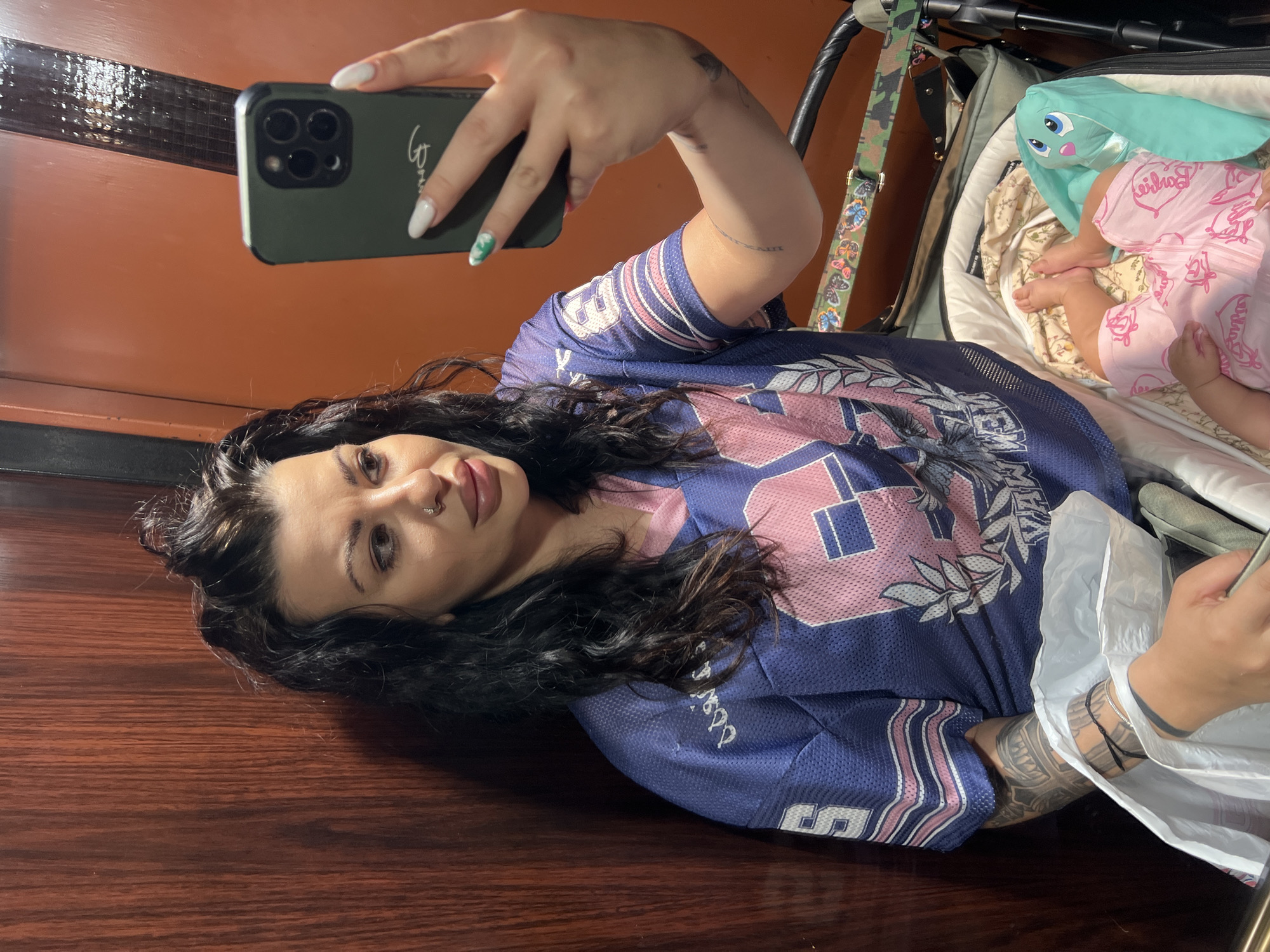 A woman is taking a selfie indoors. She is wearing a blue and pink sports jersey. To her right is a baby in a stroller, with a blue plush rabbit next to them. The background appears to be a wooden wall. It is difficult to determine if the location is in the city of Plovdiv, Bulgaria from the image alone.