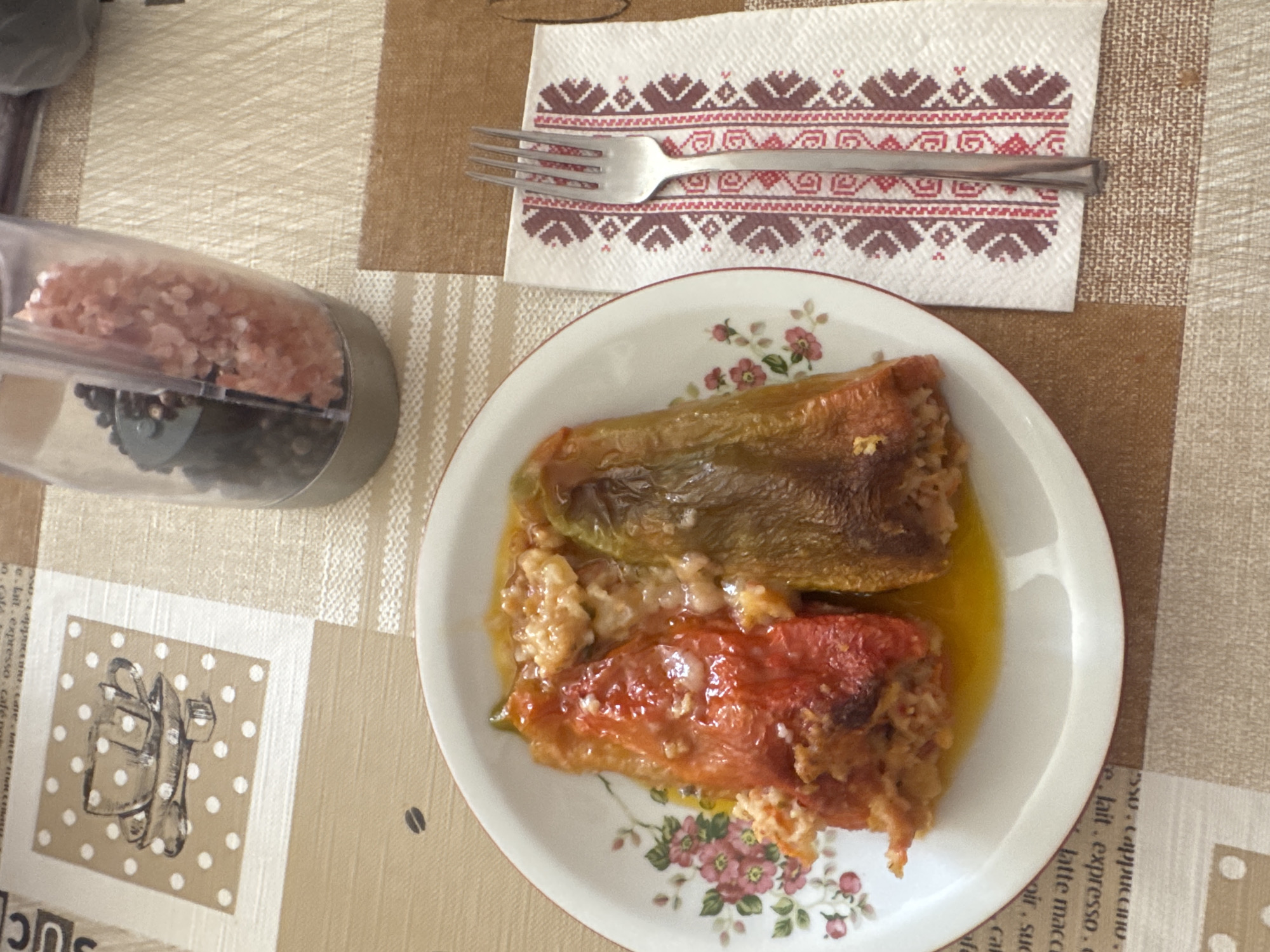 A close-up, high-angle shot shows a plate of stuffed peppers and a fork on a table. The plate contains two stuffed peppers, one red and one green, filled with a mixture of rice and meat. The peppers are in a yellow sauce. A silver fork rests on a white napkin with a red and brown embroidered pattern. To the right of the plate, a clear plastic pepper grinder filled with peppercorns and pink salt is visible. The table is covered with a brown and beige checkered tablecloth. In the background, a square placemat with a drawing of a coffee cup and dots is partially visible. The scene appears to be set indoors in a domestic environment.