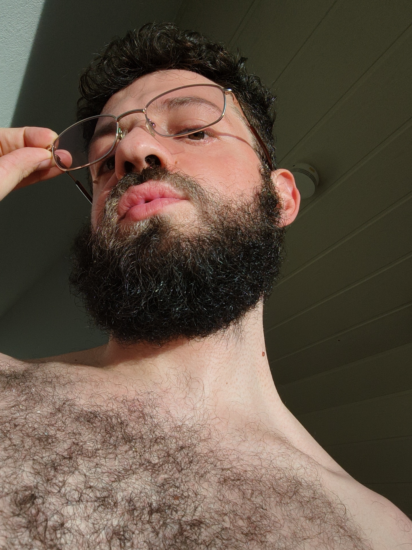 A man is shown in a low-angle, close-up shot. He has dark, curly hair, a full beard, and is wearing gold-rimmed glasses. His right hand is holding the frame of the glasses away from his face. He is shirtless, revealing chest hair and a mole on his neck. The background appears to be an interior space with a white, paneled ceiling. Light is streaming in from the side, casting shadows across the scene. There is no indication of the location in the city of Diepholz, Germany.
