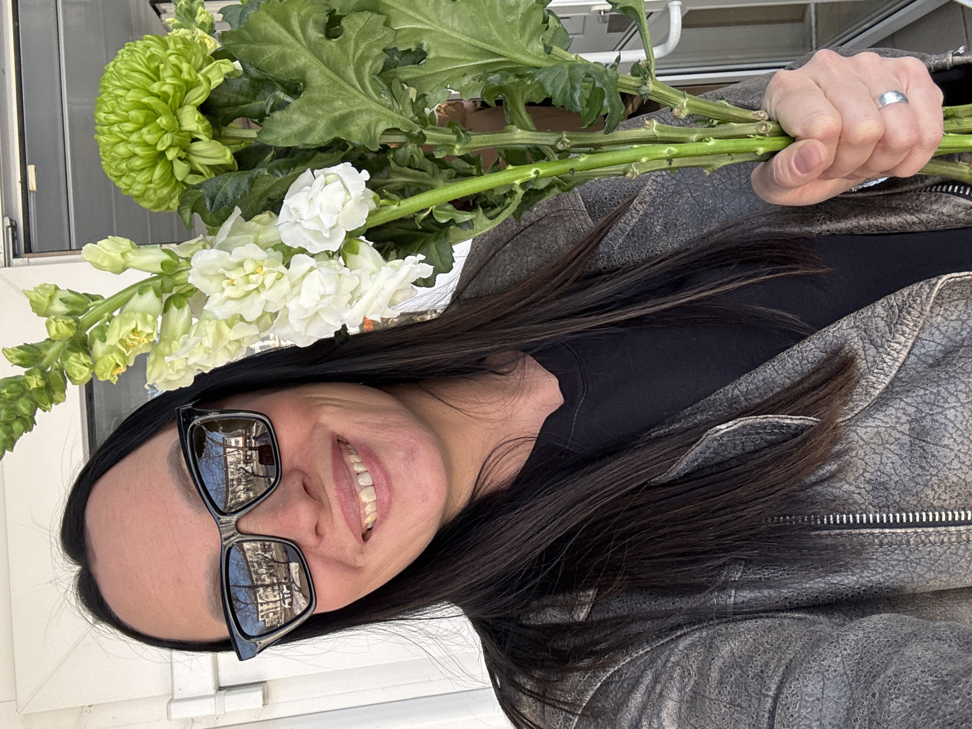 A woman with dark hair and sunglasses is holding a bouquet of flowers. She is wearing a black top and a weathered gray leather jacket with a zipper. The bouquet includes a large green pom-pom flower, white flowers, and some foliage. The reflection in her sunglasses shows a building with trees and possibly other structures. The scene seems to be outdoors, with the woman standing by a white surface. Although I cannot confirm the location is in Chaikovsky, Russia, the reflection in the glasses could provide clues if the buildings are identifiable.