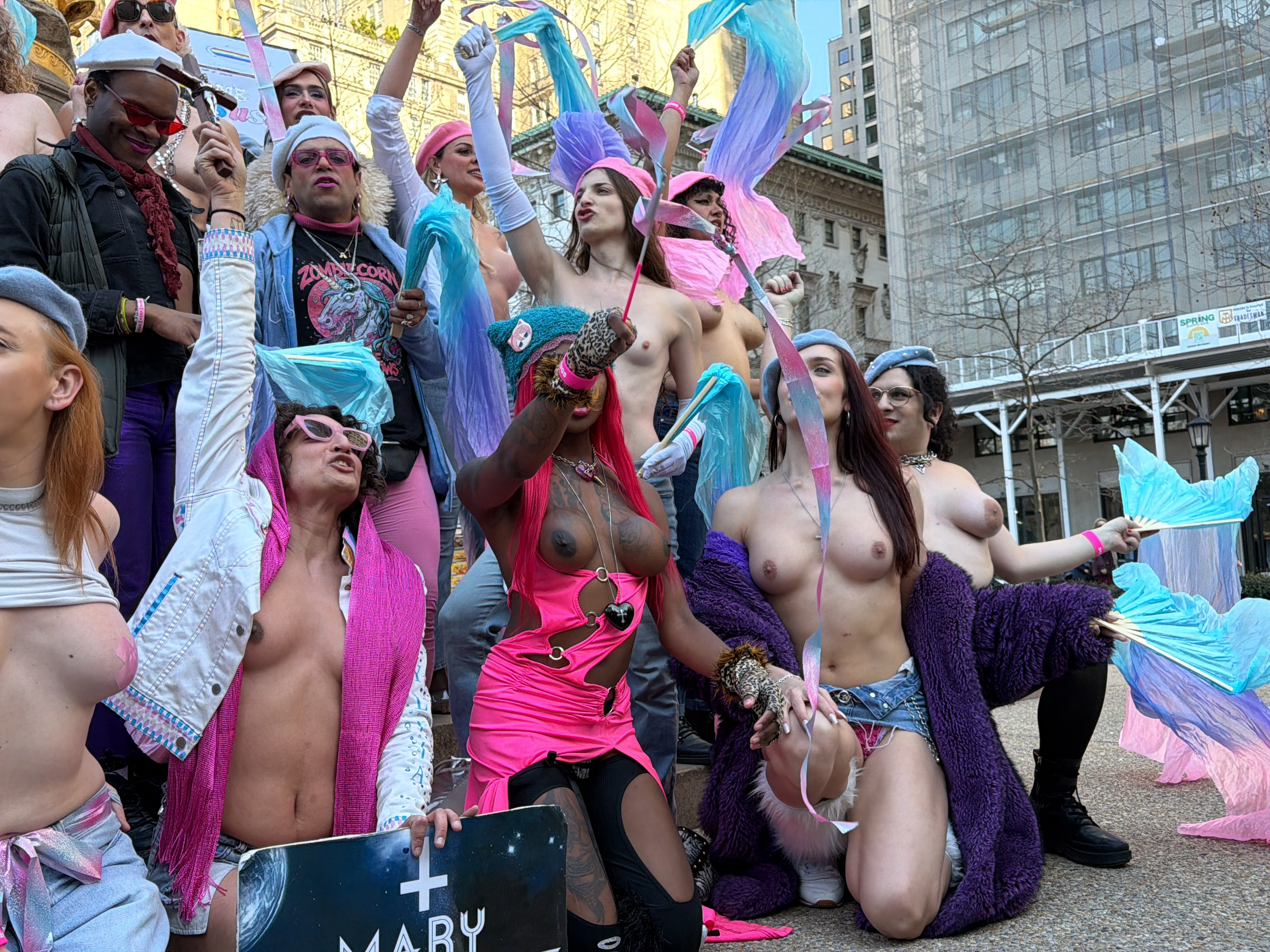 A crowd of people, mostly shirtless, pose in a city square in New York City, United States. They are of varying ages, skin tones, and genders, some holding rainbow-colored streamers and fans. Many are wearing hats and jewelry. Some are holding signs. Buildings are visible in the background. The scene appears to be a protest or demonstration.