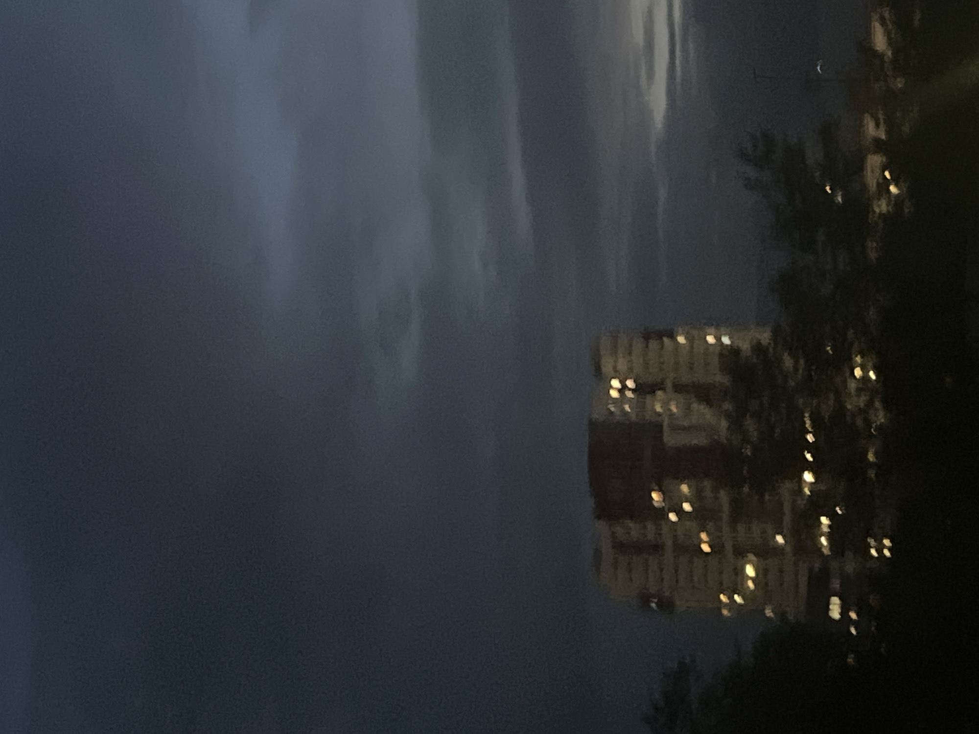 The image captures a dimly lit outdoor scene, likely taken at dusk or during the early evening, given the dark, cloudy sky and illuminated windows of a building. The main subject is a multi-story building, possibly an apartment complex, with numerous windows lit up, suggesting occupancy. The building appears to be made of concrete or a similar material, with a reddish-brown facade.
Trees with dark foliage are visible in the foreground and to the right, partially obscuring the view of the building and the sky. The sky itself is a deep, moody blue-gray, filled with swirling clouds that create a dramatic, somewhat somber atmosphere. Faint streaks of lighter gray and white in the clouds indicate the direction of the wind or variations in cloud density.
In the upper right portion of the sky, a faint crescent moon is discernible, a delicate contrast to the dark clouds. The overall setting suggests an urban or suburban environment, with the building representing residential architecture. The lighting conditions create a sense of twilight and perhaps a hint of an approaching storm due to the dark clouds. No people are visible in the frame, and there is no visible text. The image evokes a quiet, contemplative mood, possibly highlighting the transition from day to night in an overcast environment.