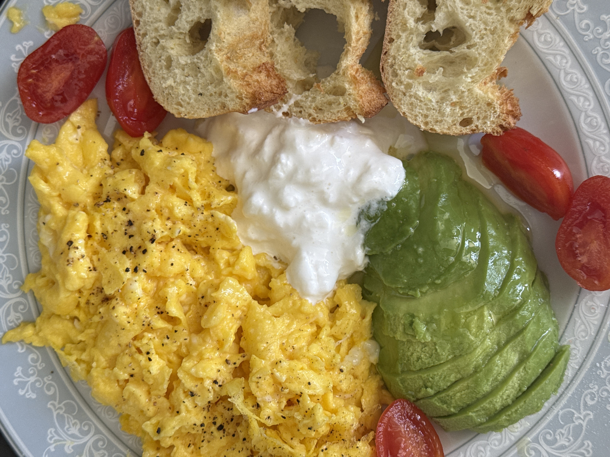 A top-down shot shows a plate of food consisting of scrambled eggs, sliced avocado, a dollop of white cream, halved cherry tomatoes, and pieces of bread. The scrambled eggs are seasoned with black pepper. The plate has a blue and white decorative pattern around the rim. There is no indication of the location being Milan, Italy.