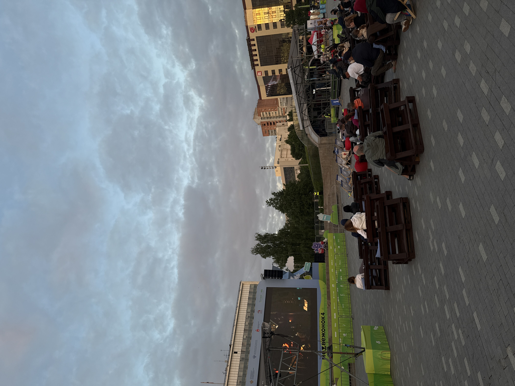The image depicts an outdoor event taking place in Perm, Russia. A large screen displaying an event called "Mozzhevelonok 4" is visible. People are seated on makeshift benches made of stacked wooden pallets, watching the screen. In the background, buildings typical of a city environment are visible, with a cloudy sky overhead. The ground is paved with interlocking gray stones. The event seems to be taking place in a public square or open area. There are green banners along a railing. Some people are wearing red hats.