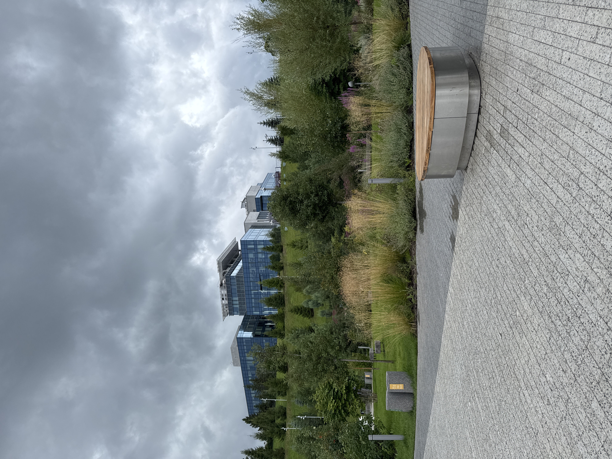 The image depicts a modern office building situated on a grassy slope in Kazan, Russia. The building features a glass facade and a flat roof with a prominent structure. In the foreground, there is a paved area with a contemporary, oval-shaped bench made of wood and metal. Beyond the paved area, a lush landscape with trees, ornamental grasses, and flowering plants is visible, leading up to the building. The sky is overcast with dramatic, dark clouds. The scene appears to be a public space or park area adjacent to the office complex. There are no people visible in the image.