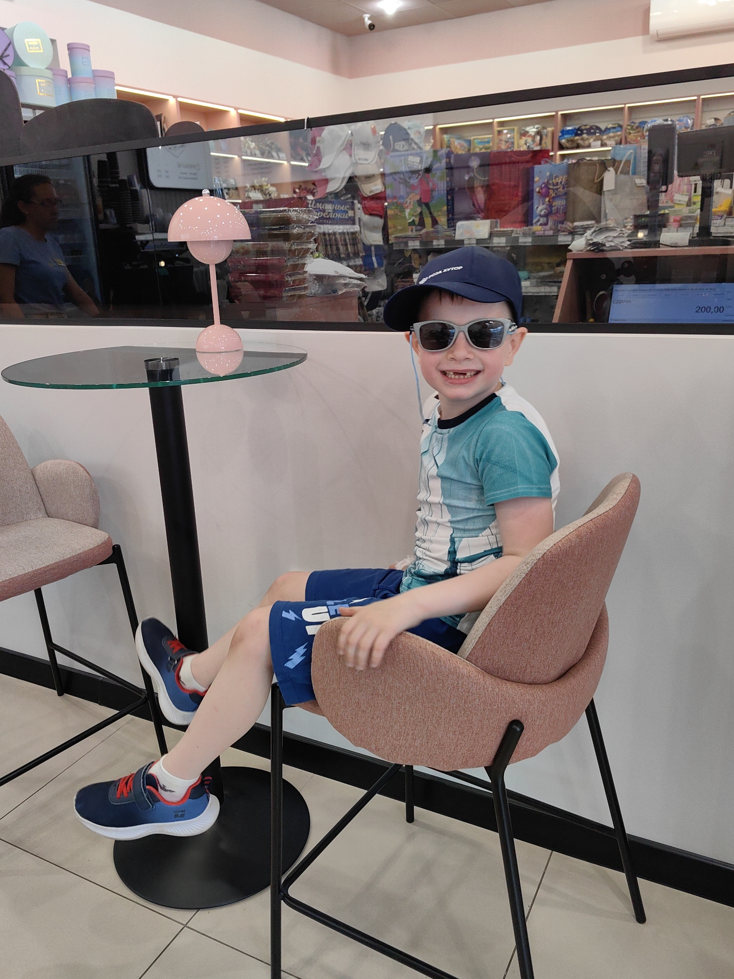 A young boy, smiling and wearing a blue baseball cap and grey sunglasses, is seated on a pinkish-orange upholstered bar stool at a glass-topped table. He wears a blue and white patterned t-shirt, blue shorts with white designs, and blue sneakers with red accents. A missing front tooth is visible as he grins towards the viewer. On the table, there is a distinct pink mushroom-shaped lamp and a small black votive or holder.
The setting appears to be an indoor commercial space, possibly a cafe or a shop, with a modern and clean aesthetic. A glass partition separates the seating area from what looks like a retail section. Behind the glass, shelves are stocked with various products, including decorative boxes in light blue and purple/pink hues. A woman wearing glasses and a blue shirt is visible as a reflection in the glass, suggesting an employee or another customer. Further back, a cash register or monitor displays "200,00", likely indicating a price.
Visible Cyrillic text includes "РОБА ХУТОР" on the boy's cap, and partially legible words like "Игрушки" (Toys) and "подарки" (gifts) behind the glass, along with "СДАЧА". The overall impression is cheerful and relaxed. The scene is situated in Sirius, Russia, as indicated by the context.