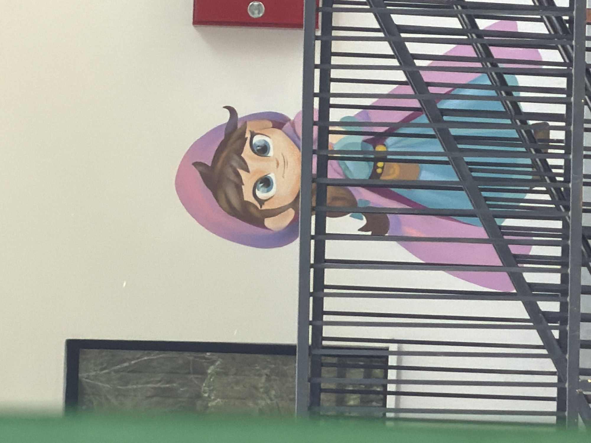 A mural depicting a cartoon character with brown hair and blue eyes, wearing a pink hooded outfit, is painted on a white wall. The mural is partially obstructed by a dark gray metal railing with horizontal bars. A black framed object or architectural feature is located below and to the left of the mural. A red object is visible above the mural. The scene is observed from a slightly elevated perspective. The image was captured in Tbilisi, Georgia.