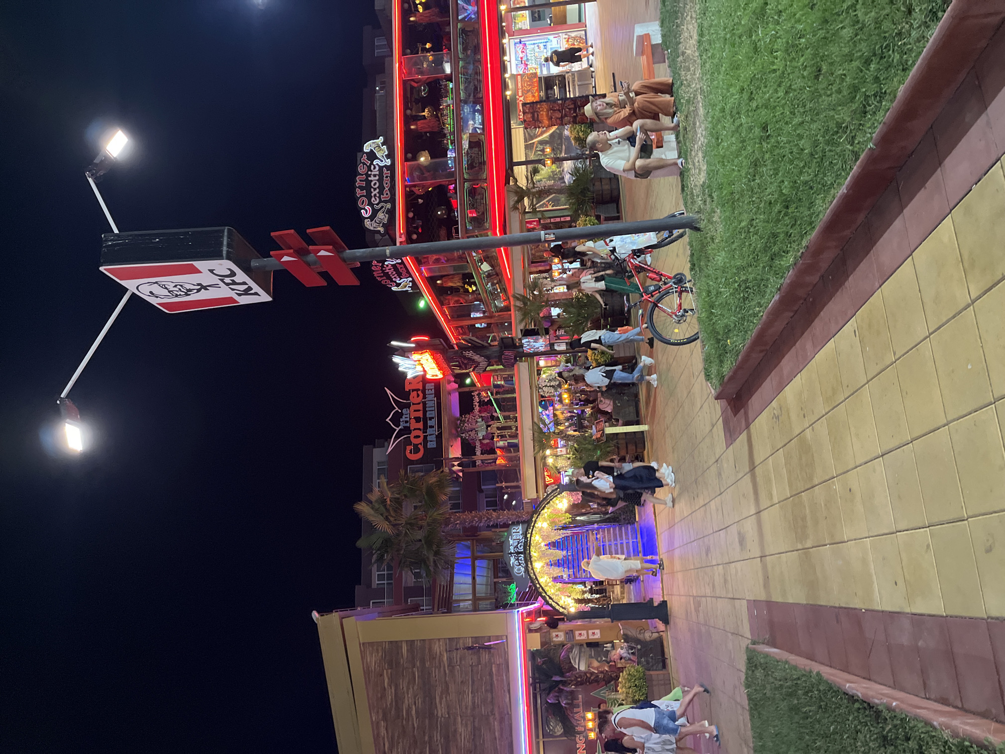 The image depicts a bustling street scene in Nesebar, Bulgaria at night. A KFC sign is prominently displayed on a lamppost, casting a bright light on the surrounding area. Several buildings line the street, adorned with neon signs and colorful lights, indicating a vibrant entertainment district. People are seen walking along the paved walkway, some are sitting on a bench, and others are interacting with each other. A red bicycle is parked near the lamppost. The overall atmosphere is lively and energetic, with a mix of commercial establishments and pedestrian activity.