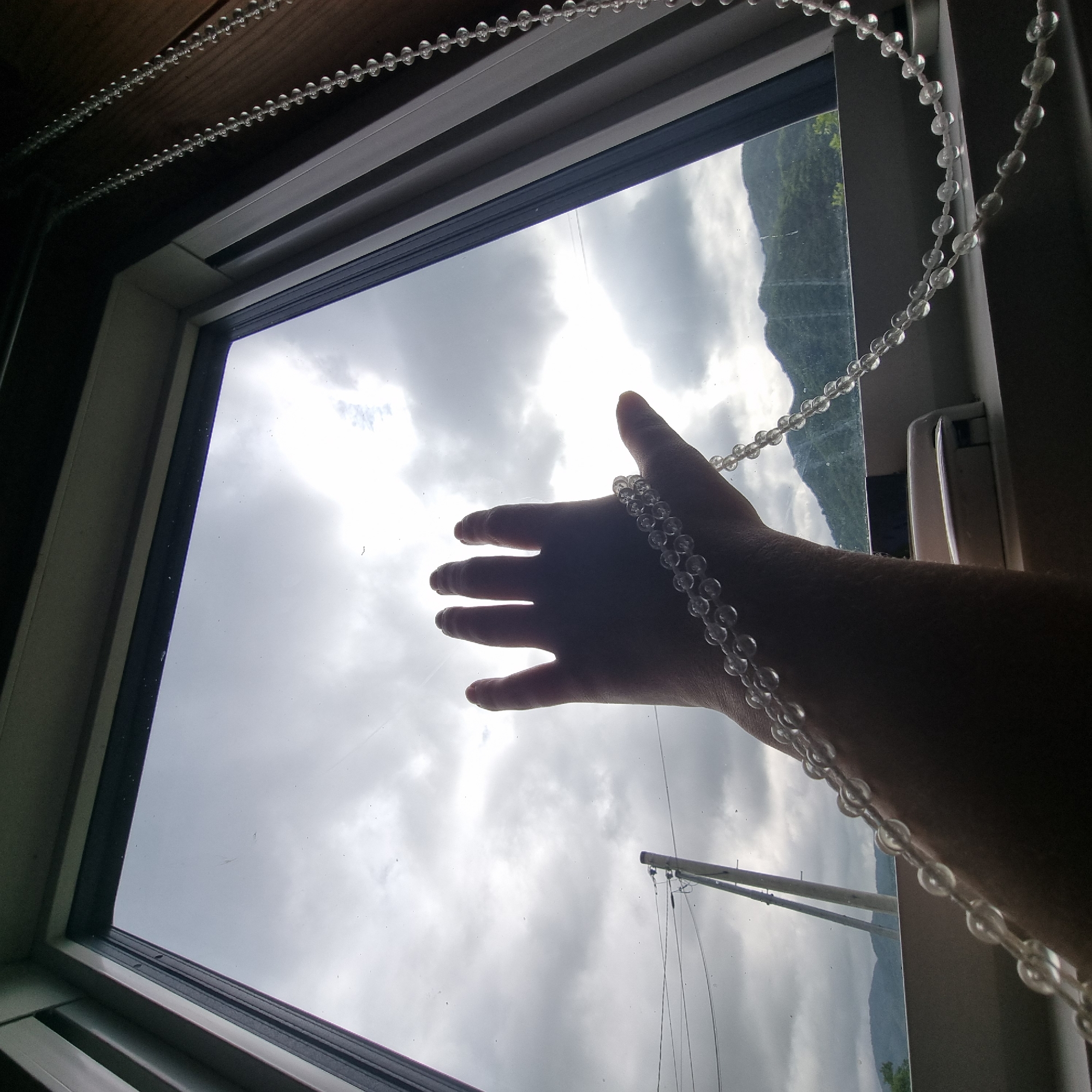 A person's outstretched hand is silhouetted against a cloudy sky as seen through a window in Dunnae-myeon, South Korea. The hand is adorned with a string of translucent beads that also drapes across the arm. Outside the window, a utility pole with electrical wires is visible, along with green, treed mountains under a gray, overcast sky. The window frame is white, and a white blind pull cord with clear beads runs along the right side of the frame. The scene appears to be viewed from inside a room, looking outwards.