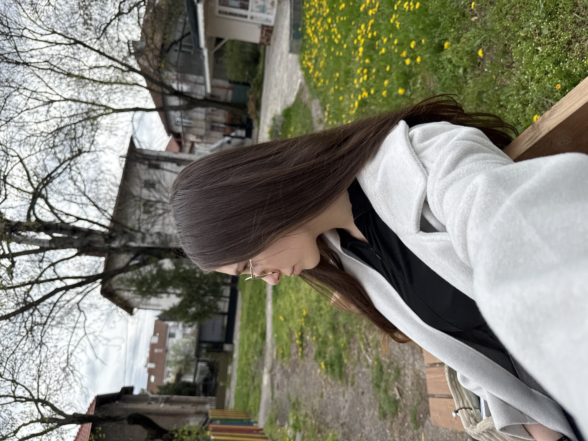 A young person with long, dark brown hair and gold-rimmed glasses is seated outdoors on a wooden bench, viewed in profile. They are looking downwards, appearing contemplative or focused. They wear a black top beneath a light grey or white textured jacket or coat.
The setting is a green area, likely a park or garden, with patchy grass and numerous small yellow flowers, resembling dandelions, covering the ground. A dirt or gravel path is also visible. In the background, several trees with sparse foliage or bare branches suggest early spring. Beyond the trees, residential buildings with varying architectural styles and visible roofs, some with red tiles, line the horizon under an overcast sky. The overall impression is a cloudy daytime scene in a suburban or urban green space in Sofia, Bulgaria. No visible text is present.