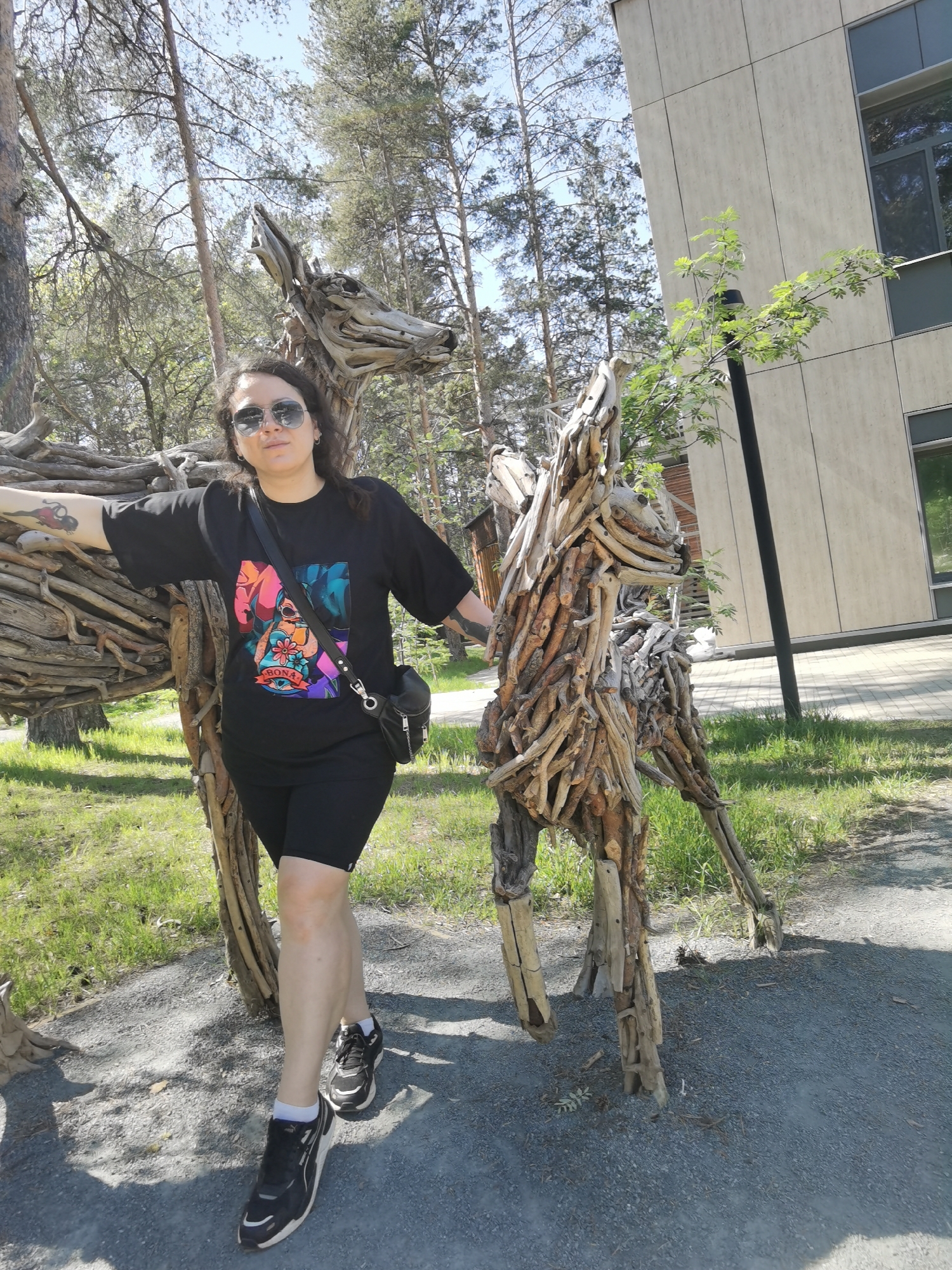 A woman with sunglasses and a black outfit stands outdoors next to two wooden sculptures of animals, likely deer or horses. The sculptures are made of sticks and branches. The woman is touching one of the sculptures with her left hand. She is wearing black sneakers with white soles. Behind the sculptures, there is grass, trees, and a modern building with windows and a light-colored facade. The ground is covered with gravel. The location is Perm, Russia.