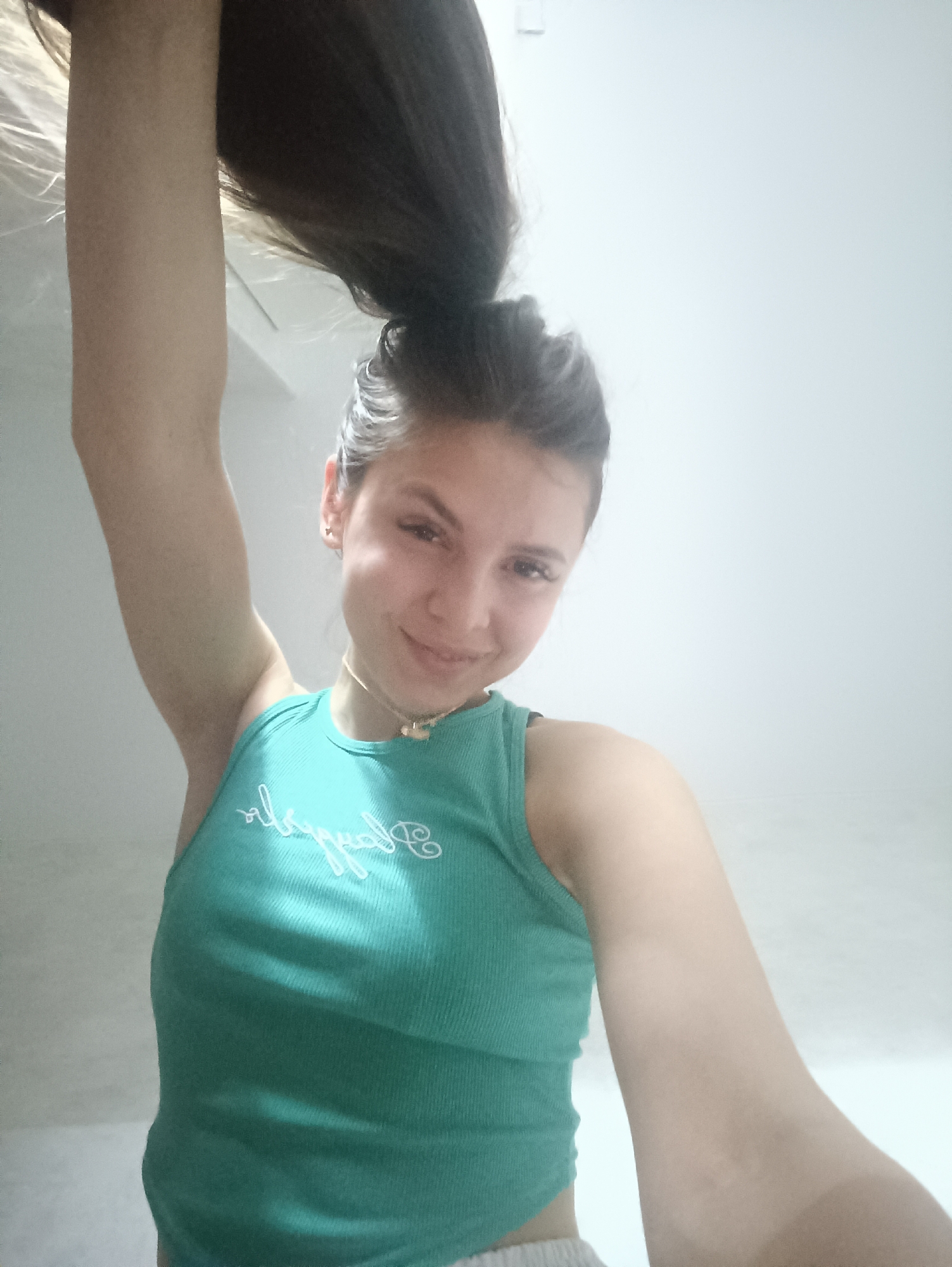 A woman is holding her dark brown hair up above her head. She has fair skin, dark brown eyes, and a gold necklace. She is wearing a light green ribbed tank top. The background is a plain white wall.